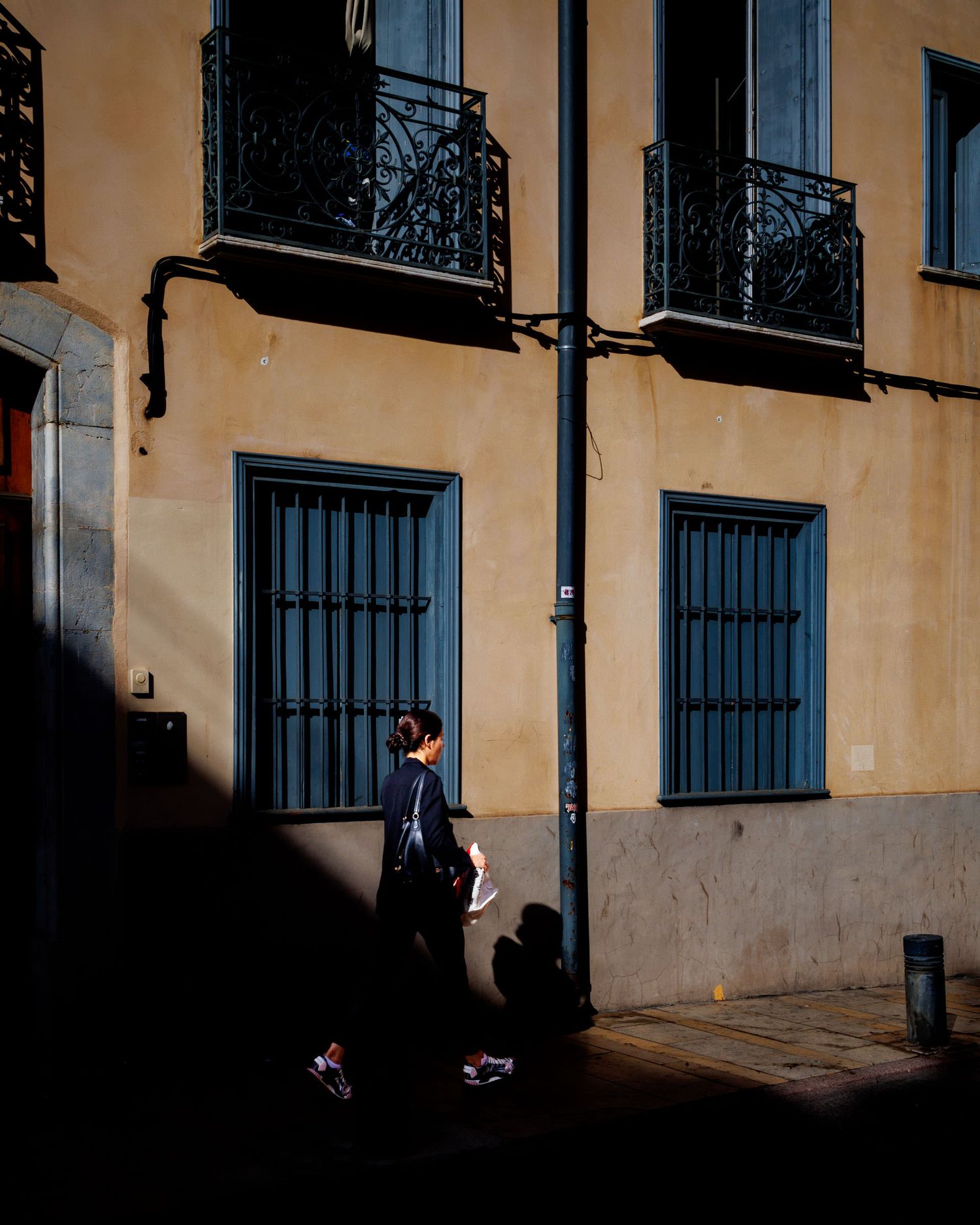 Person walking past a sunlit yellow building with barred windows and ornate iron balconies.
