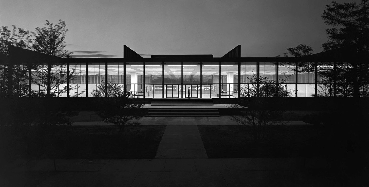 Black and white nighttime photo of a modernist glass building illuminated from within, surrounded by trees and a walkway.