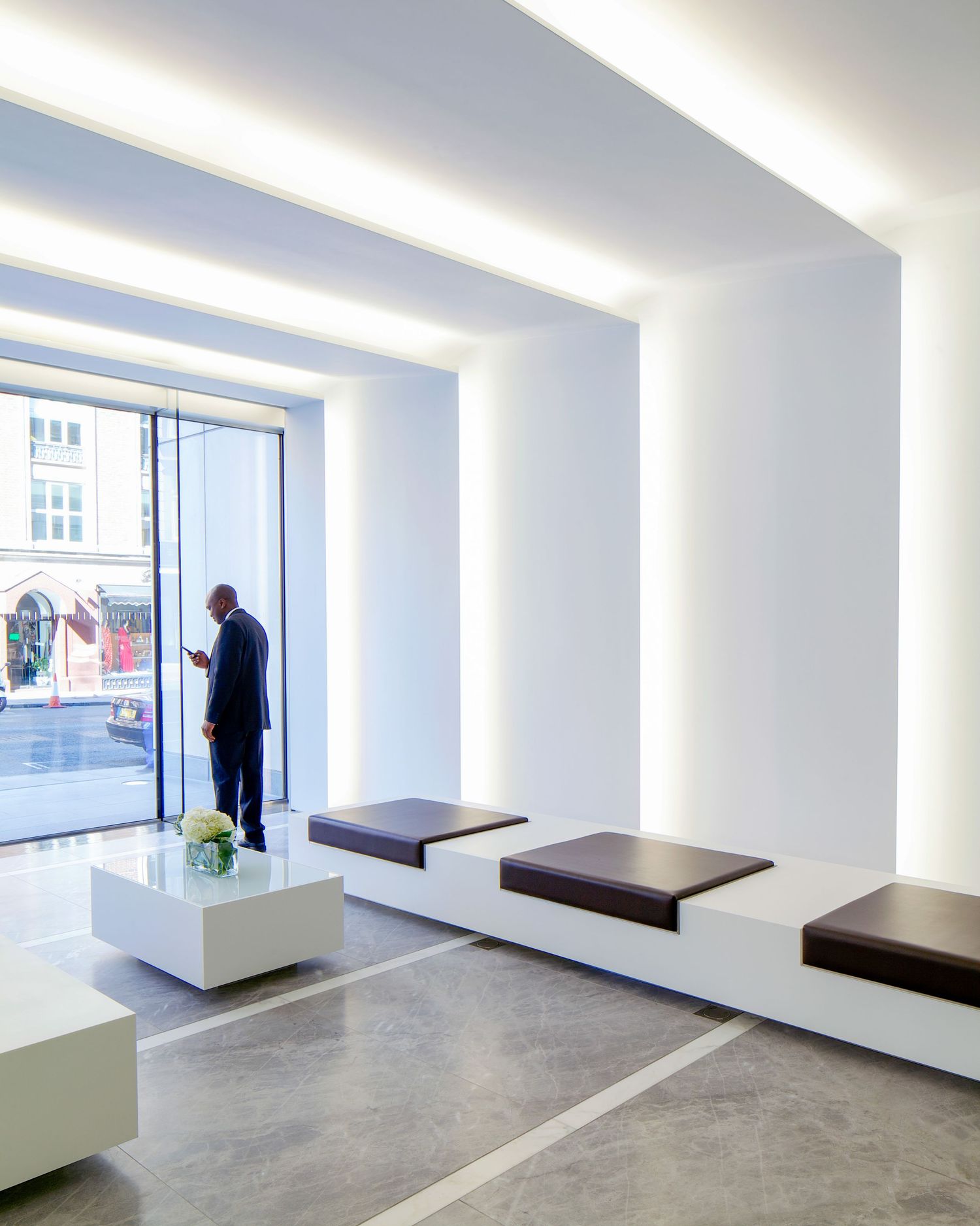Modern minimalist lobby with white walls, recessed lighting, dark cushioned benches, and a man standing by a glass entrance.