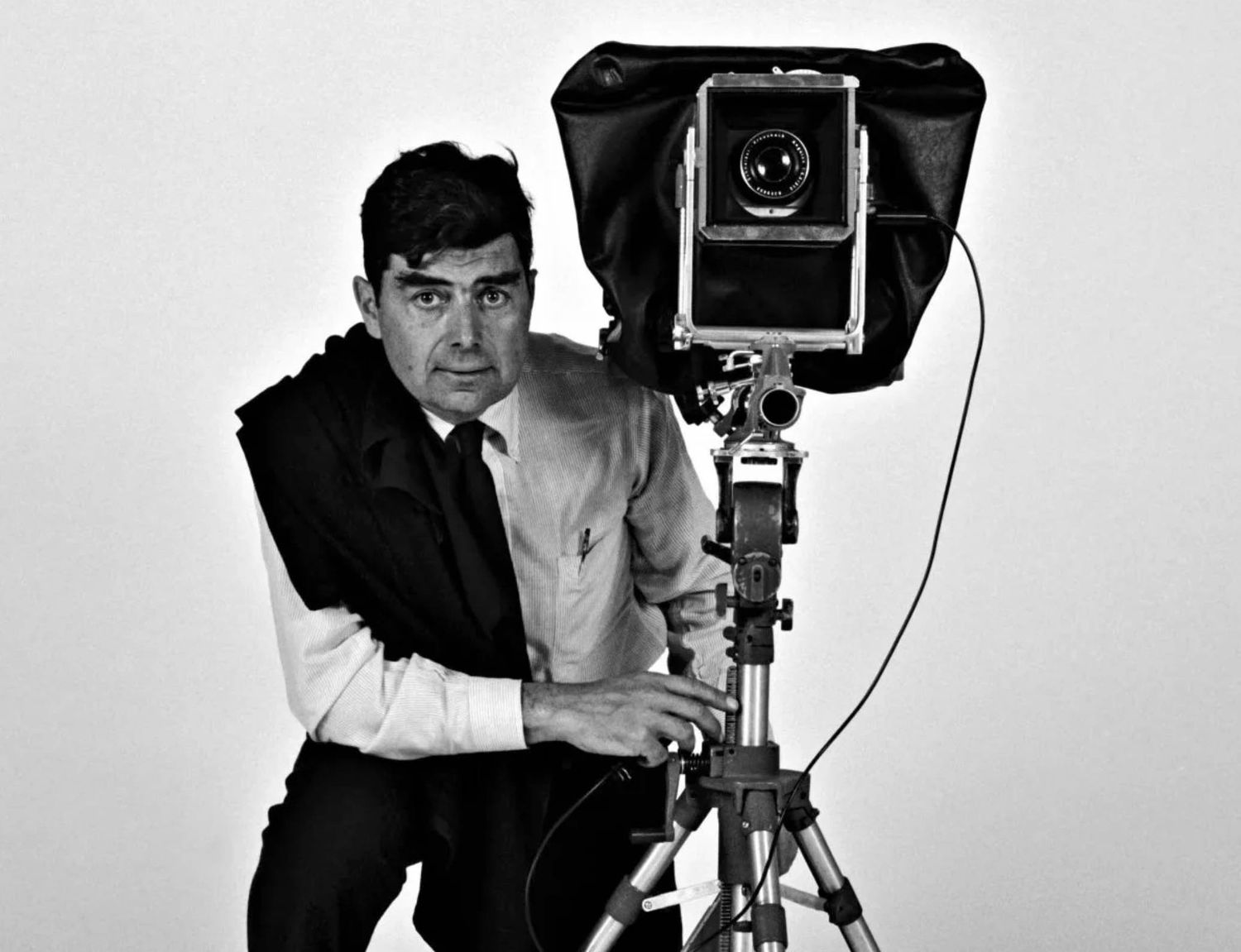 Black and white photo of a man posing beside a large vintage camera on a tripod in a studio setting.