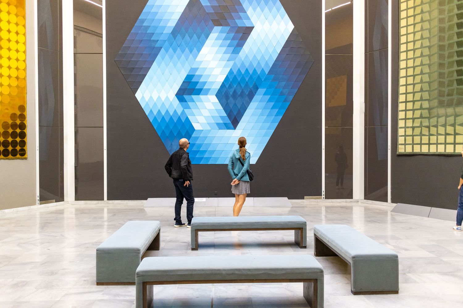 Two visitors admire a large blue geometric op-art mural in a modern gallery with benches and colorful abstract artworks.
