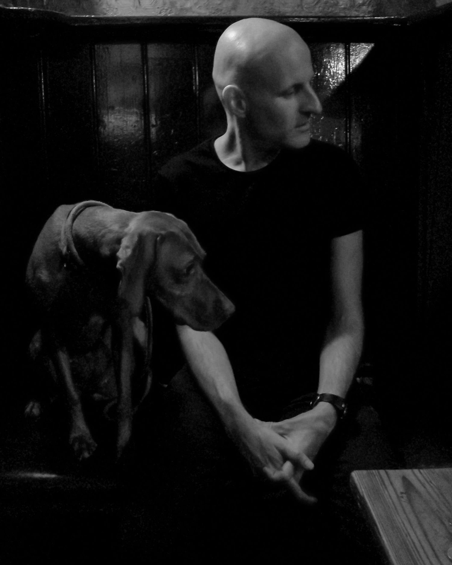 Black and white portrait of bald man in black t-shirt sitting beside a Vizsla dog.