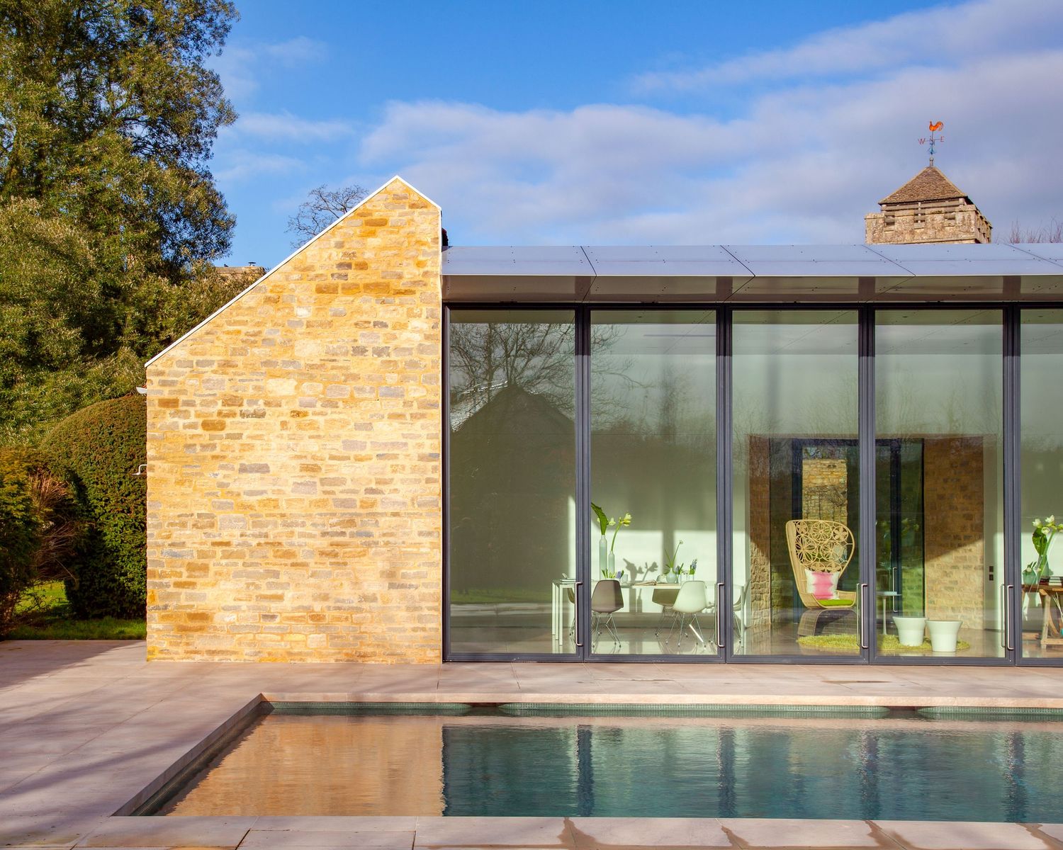 Modern glass pool house with limestone brick wall beside a sleek outdoor swimming pool set in a lush garden.