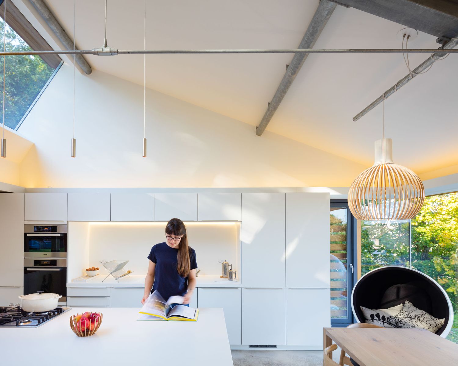 Modern white kitchen with vaulted ceiling, pendant lights, and a woman reading a cookbook at a large island.