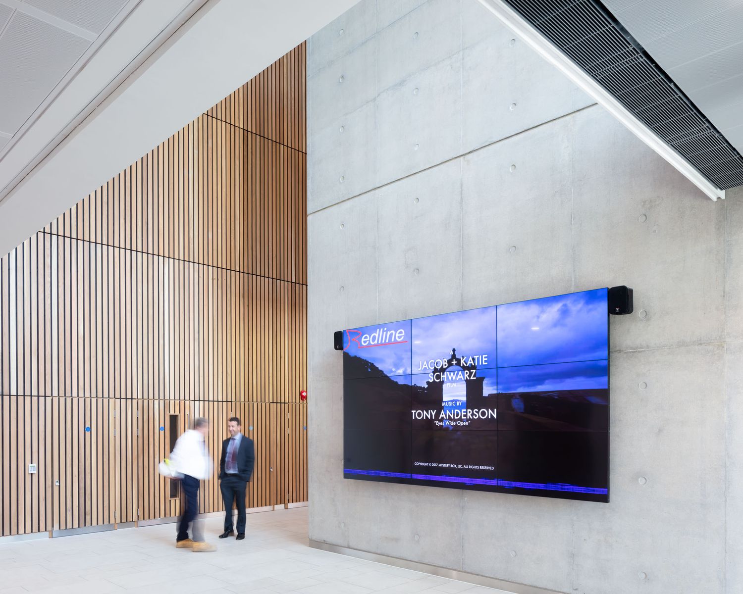 Two people stand before a large digital display screen in a modern concrete and wood lobby.