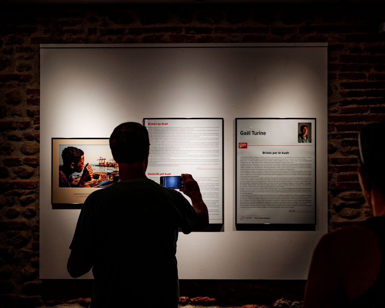 Silhouetted person photographing exhibition panels with artwork and text on a brick wall gallery.