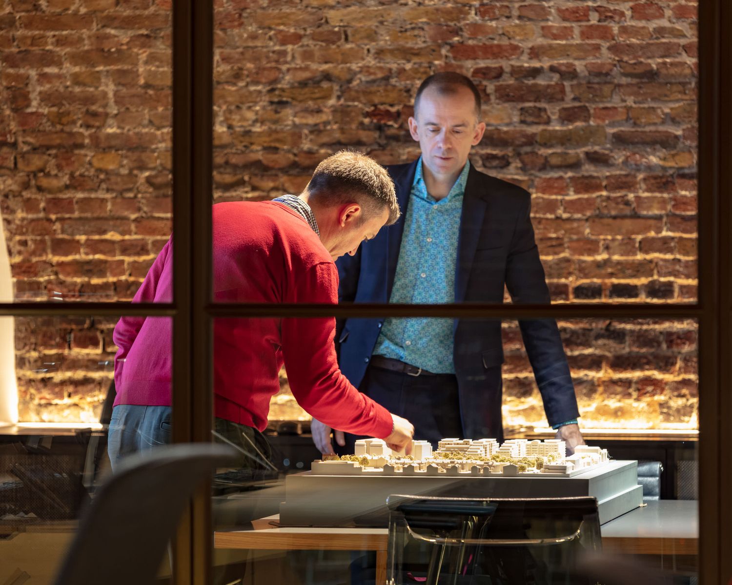 Two men examine architectural models on a table in a modern office with exposed brick walls.