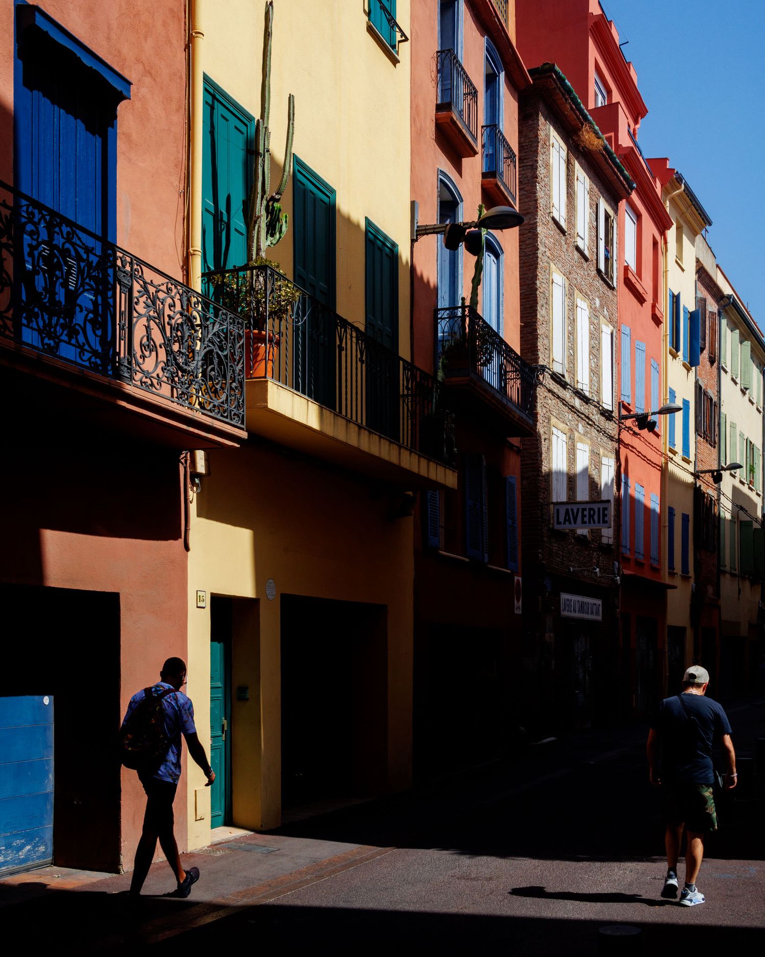 Two people walking on a sunny street lined with colorful Mediterranean-style buildings with balconies.