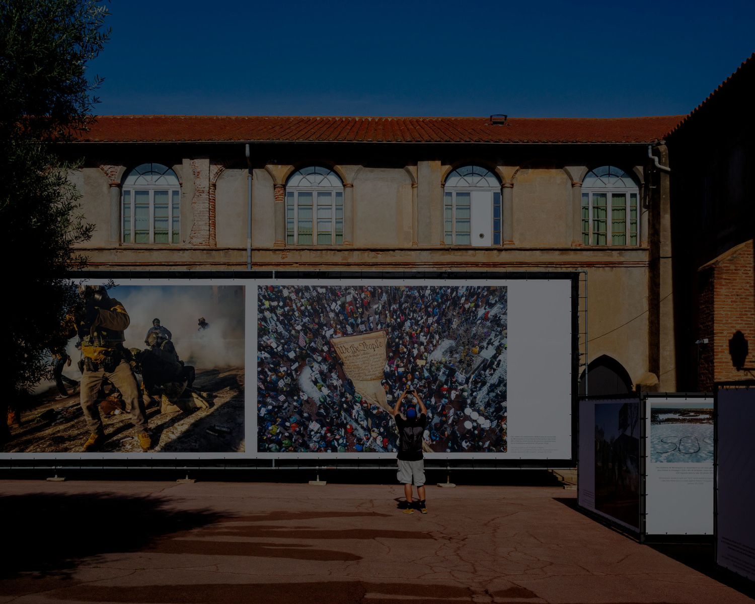 Person viewing large outdoor photography exhibition panels mounted on a historic building facade in a sunny courtyard.