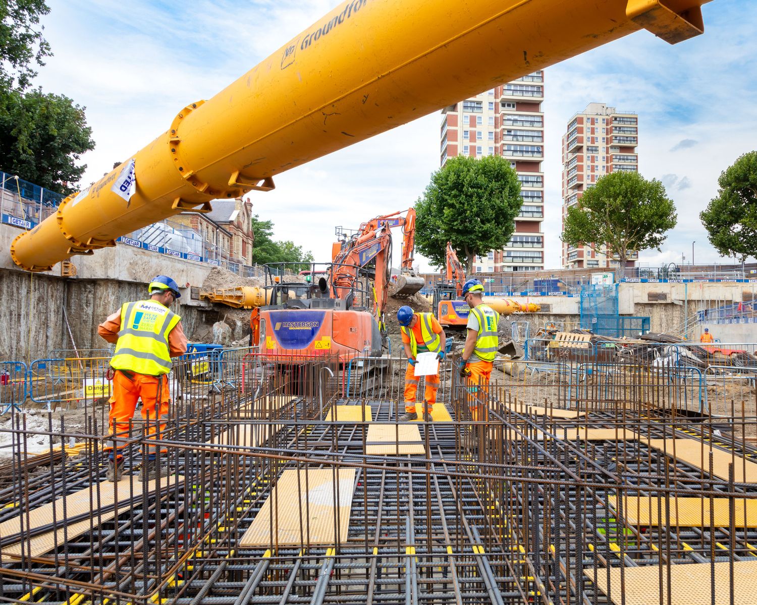 Construction workers in orange safety gear work on a large reinforced concrete foundation at an urban building site.