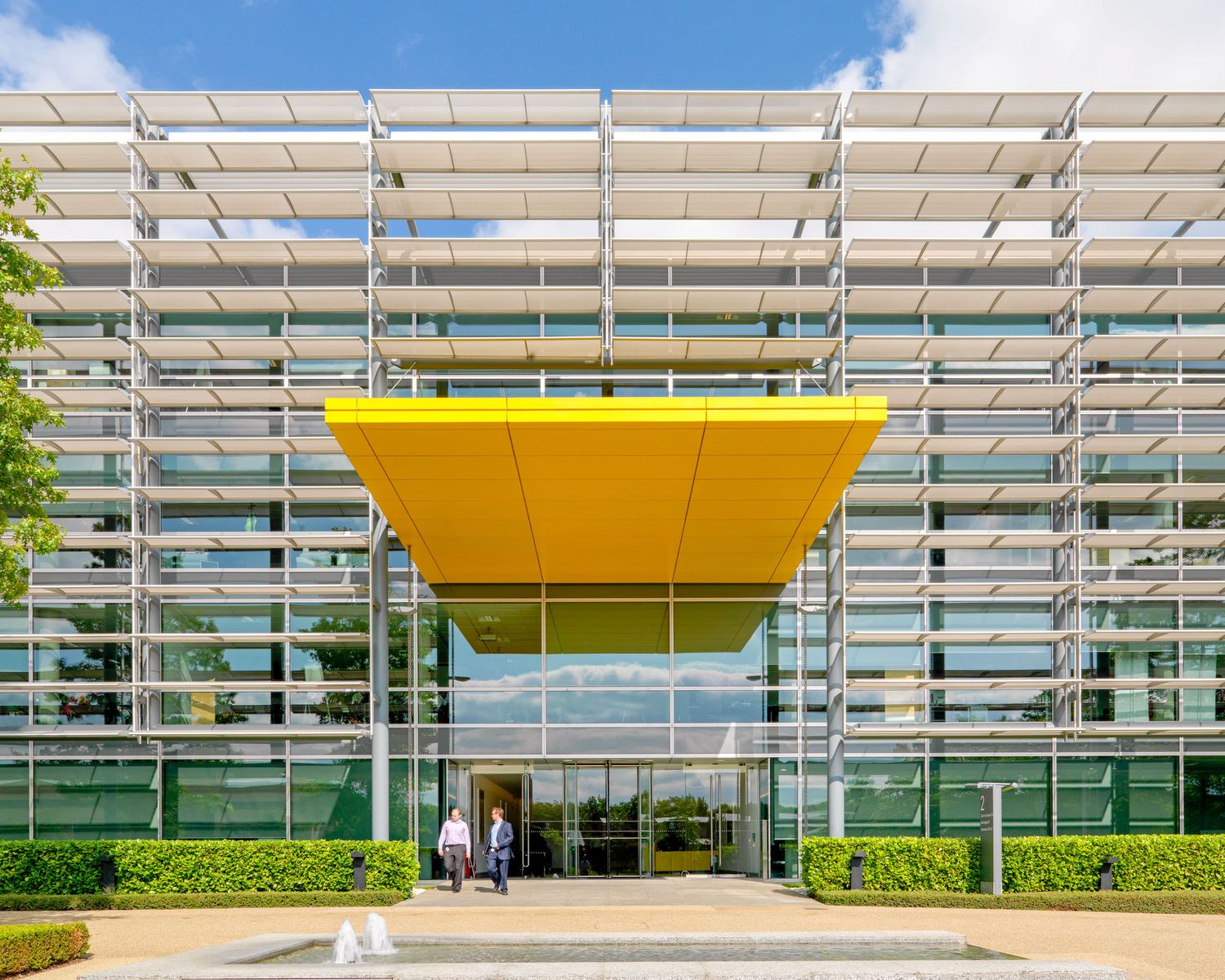 Modern glass office building with striking yellow canopy entrance, surrounded by manicured hedges under a blue sky.