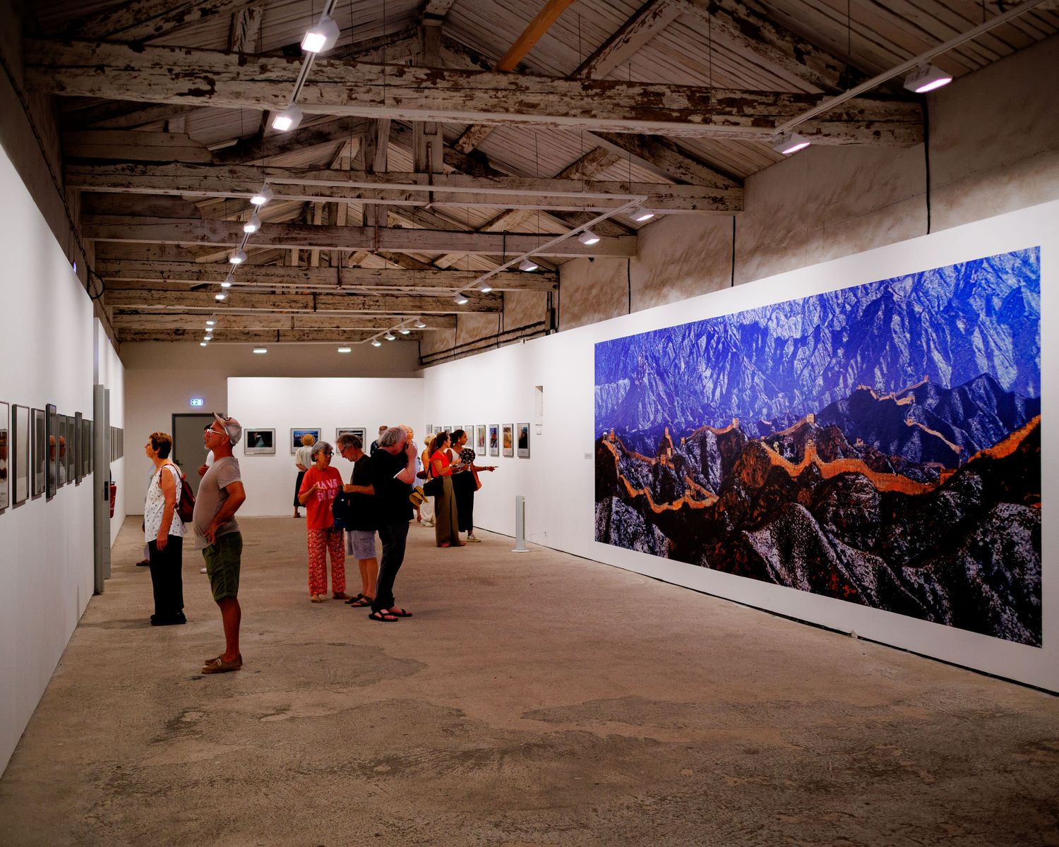 Visitors viewing large-scale photography exhibitions on white walls inside a rustic industrial gallery space.