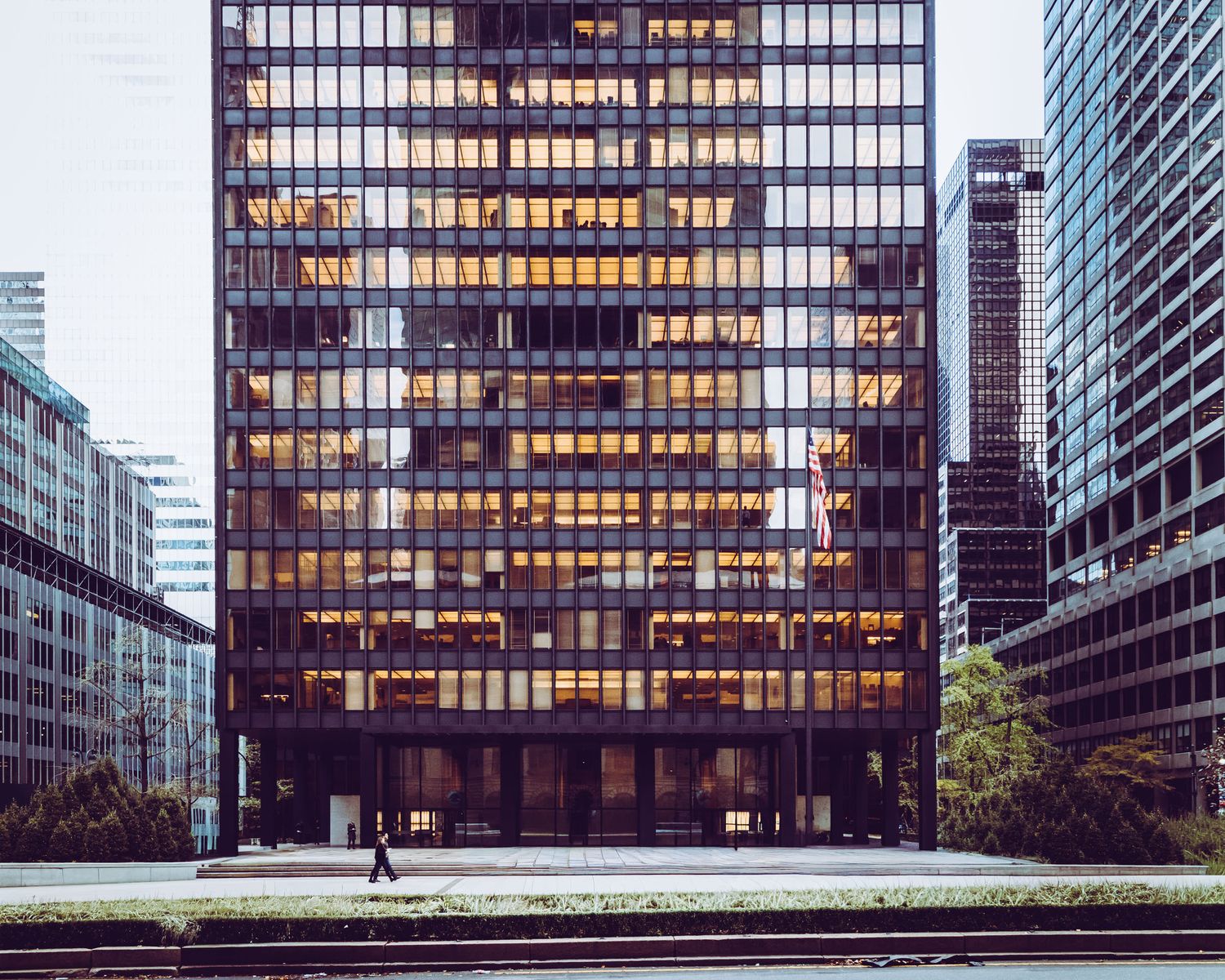 A modernist glass office tower glows with warm light against an urban cityscape at dusk.