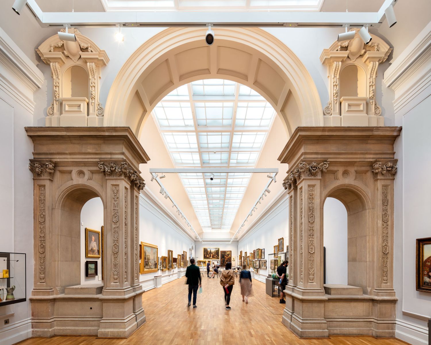 Grand museum gallery with ornate stone archway, skylight ceiling, and visitors viewing classic paintings on white walls.