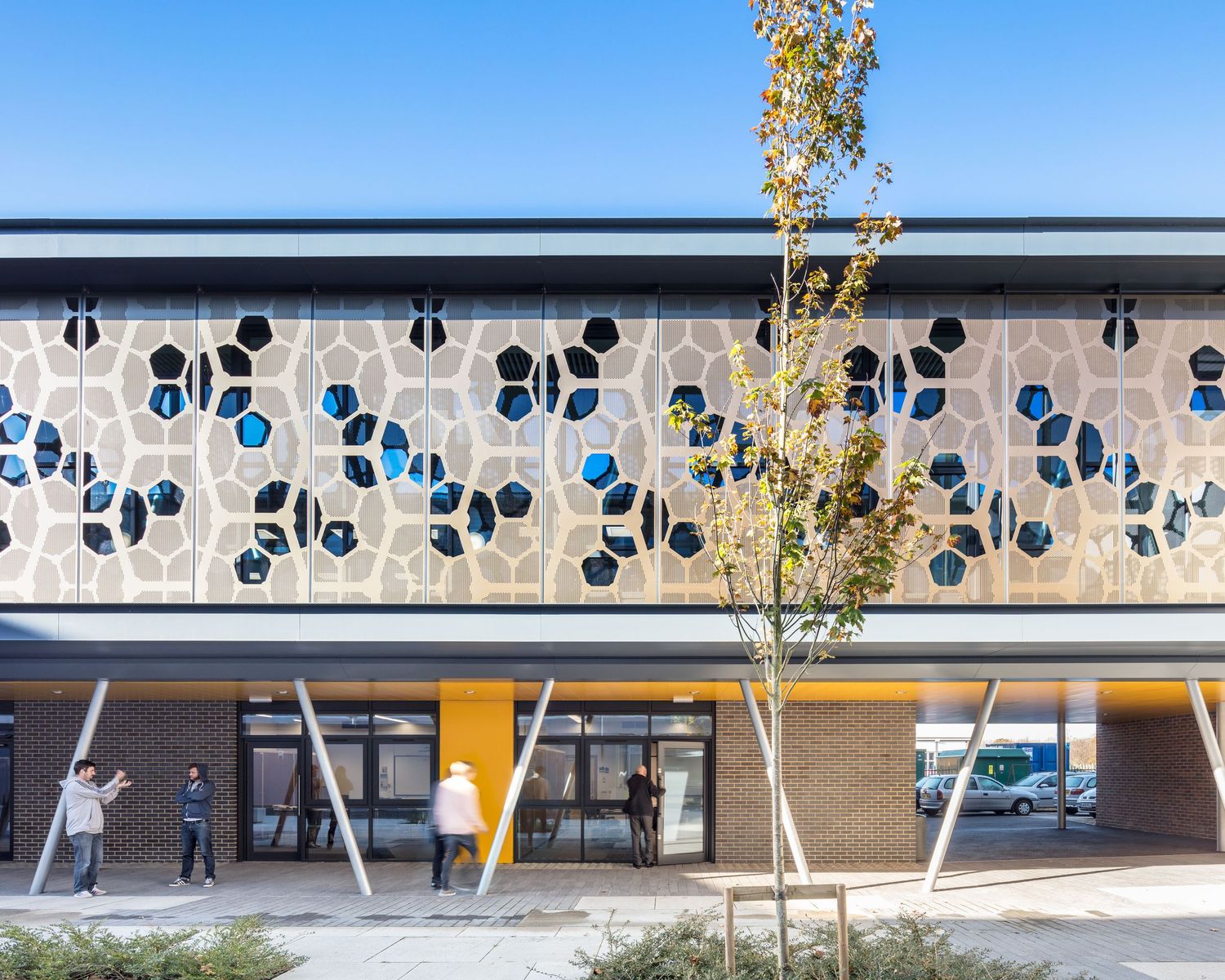 Modern building facade with decorative hexagonal patterns and blue accents, featuring yellow pillars and people walking outside.