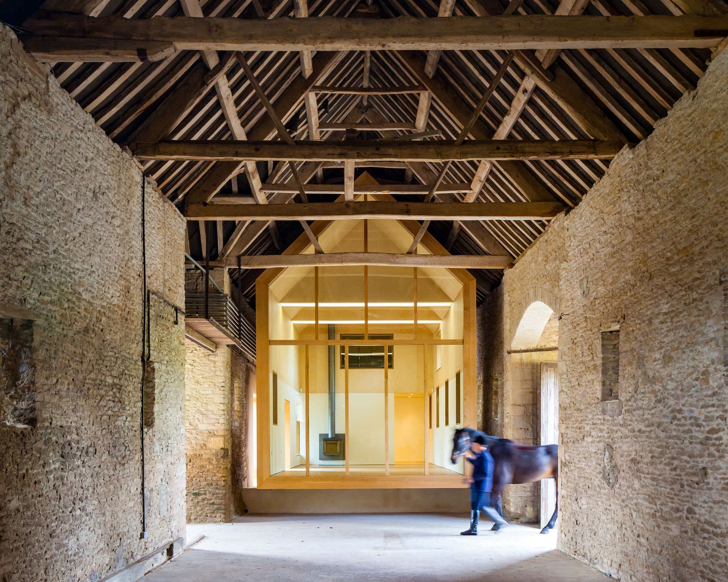 Historic stone barn interior with wooden beam roof structure and person leading a horse through arched passageway.