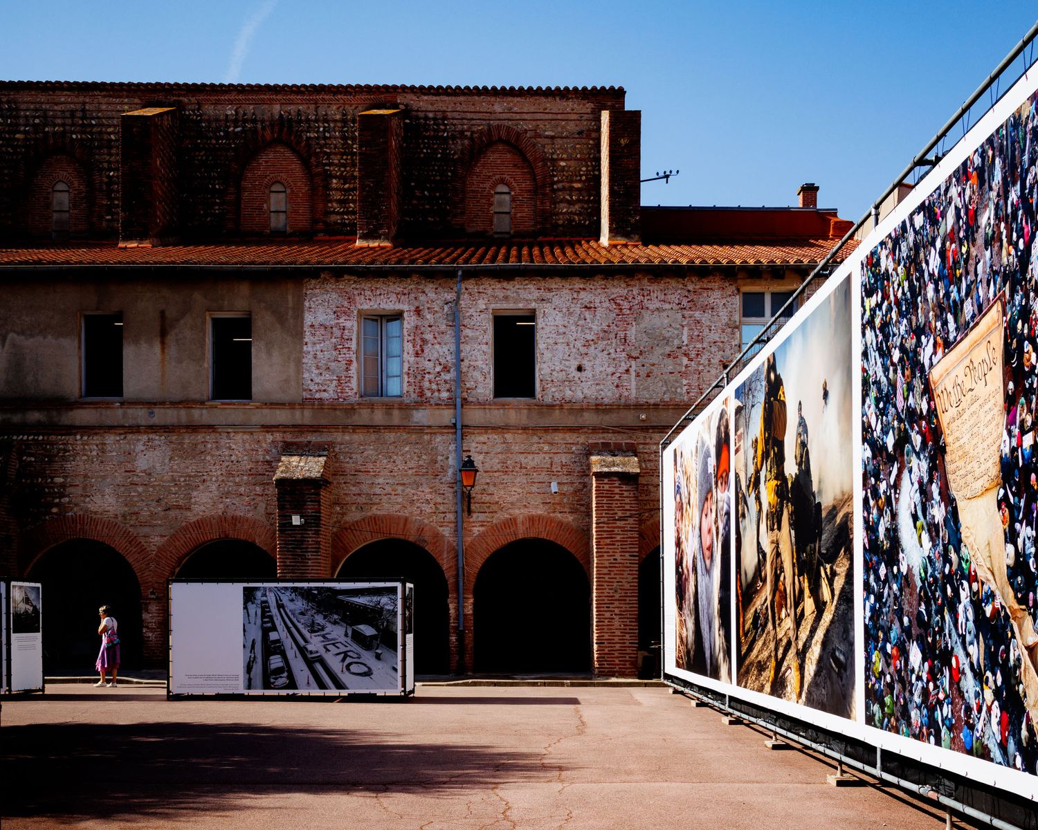 Large-scale photography exhibition panels displayed in a historic brick courtyard under a clear blue sky.