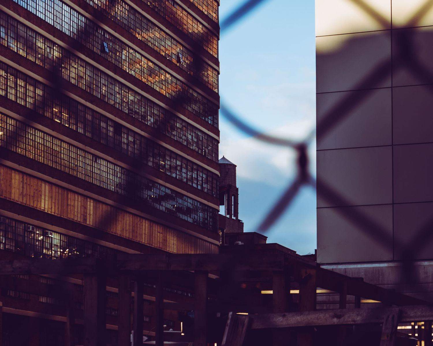 Urban industrial buildings viewed through a chain-link fence at dusk with blue sky backdrop.