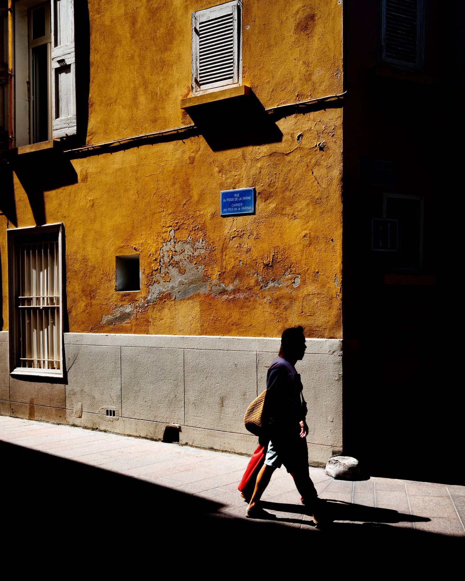 Pedestrian walking past a sunlit ochre-colored building with weathered walls and shuttered windows on a narrow street.