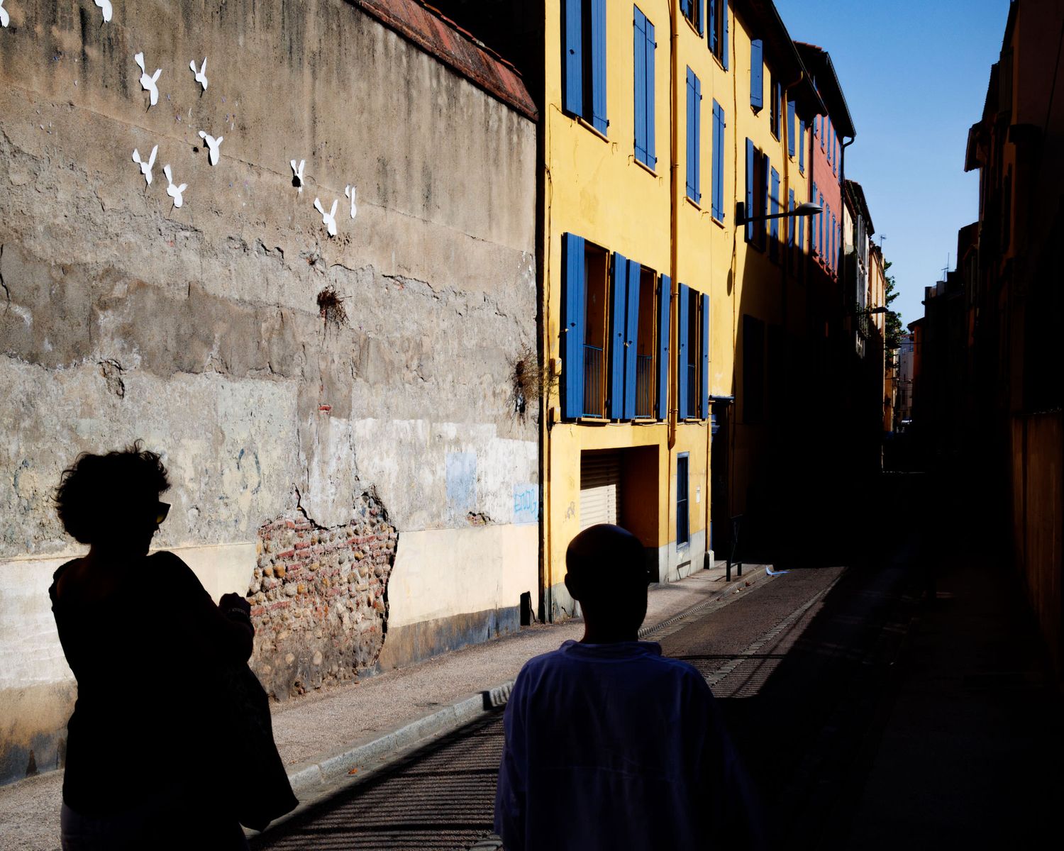 Two silhouetted figures on a cobblestone street beside a wall with bird mural and colorful buildings.