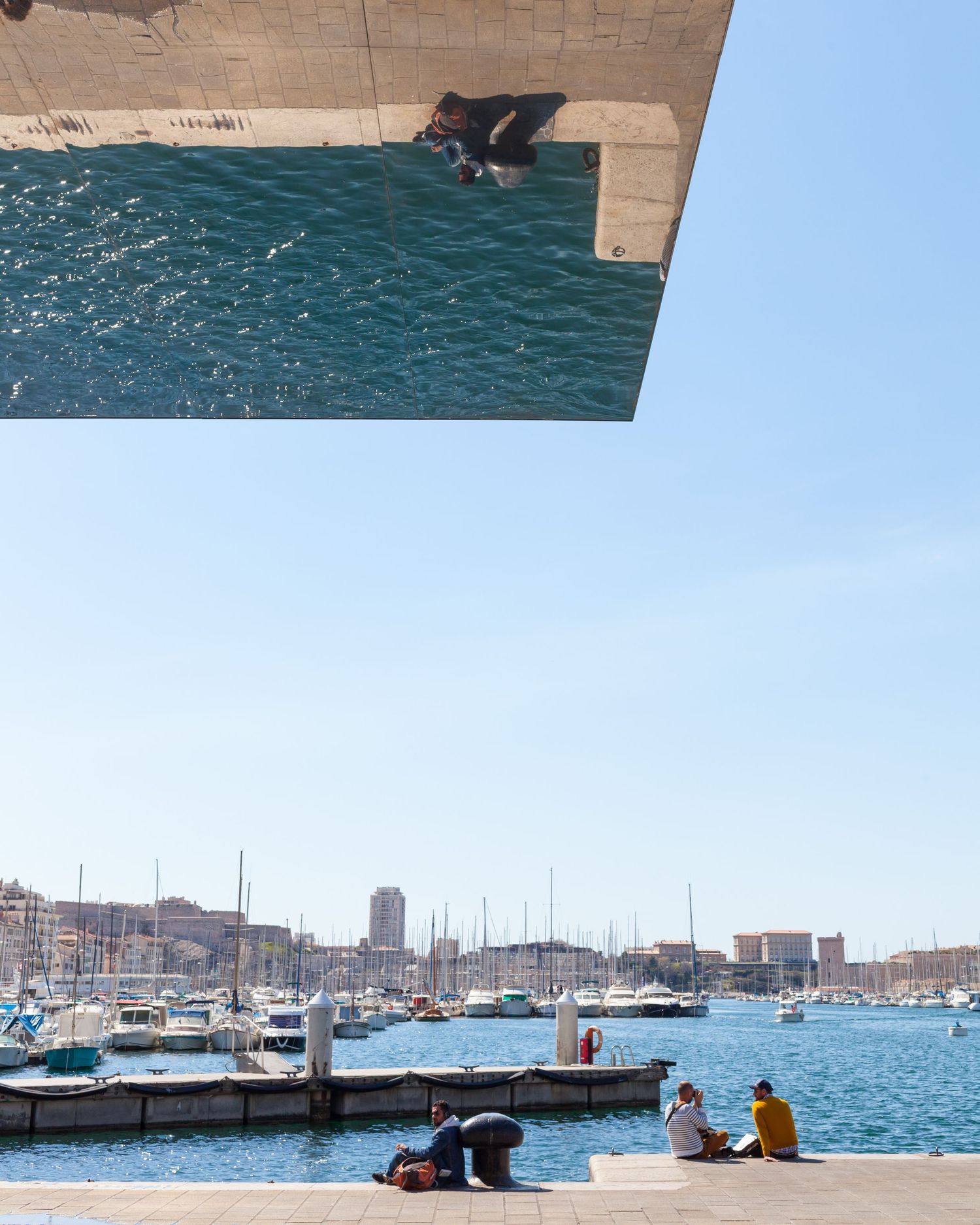 A creative split photo of Marseille harbor with boats, people relaxing by the waterfront, and a mirrored reflection overhead.
