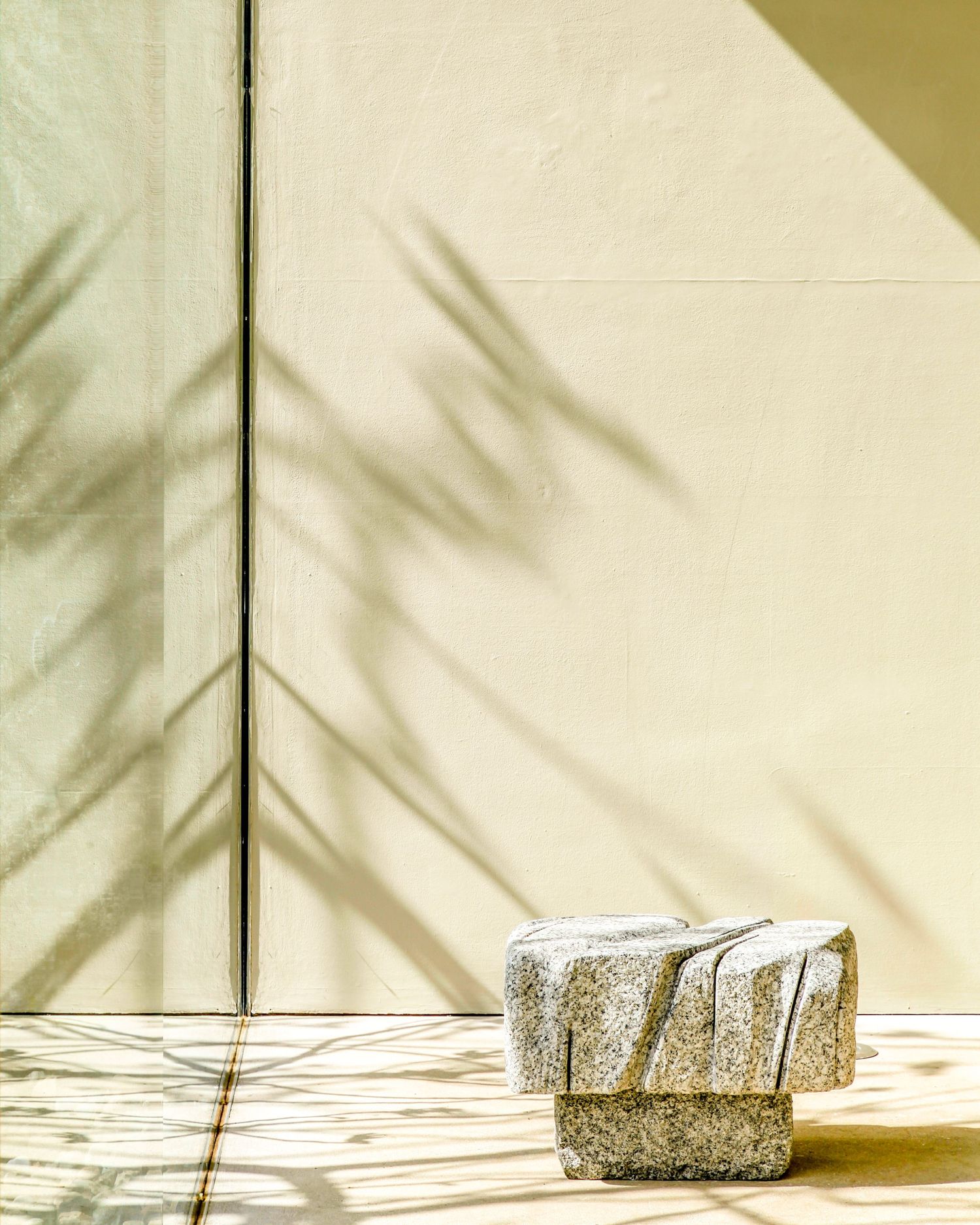 Granite stone sculpture sits on sunlit floor with palm leaf shadows on cream wall backdrop.
