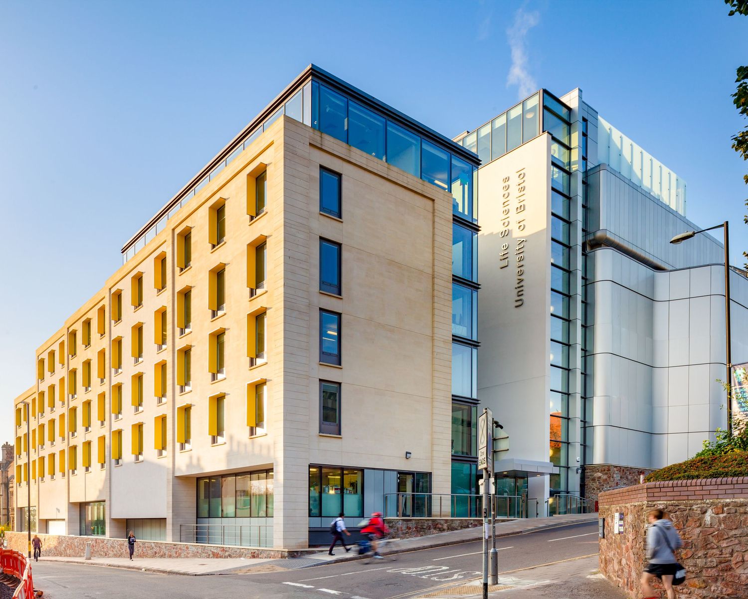 Modern multi-story hotel building with glass and stone facade on a sunny urban street corner.