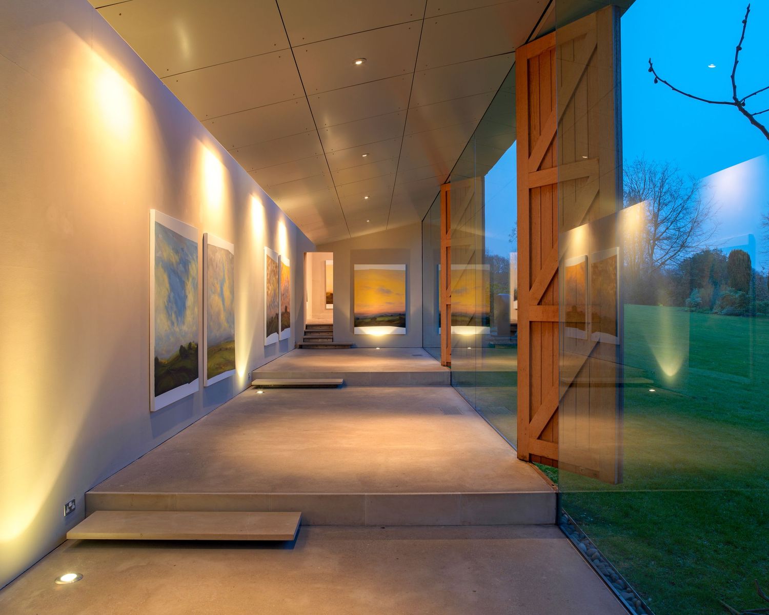 Modern art gallery corridor with warm uplighting, glass walls, and lush green lawn views at dusk.