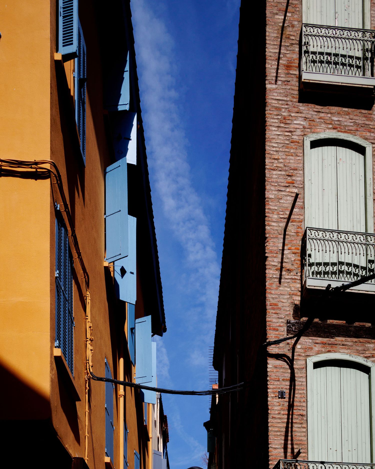 Narrow alley between colorful yellow and brick buildings with blue shutters against a bright blue sky.