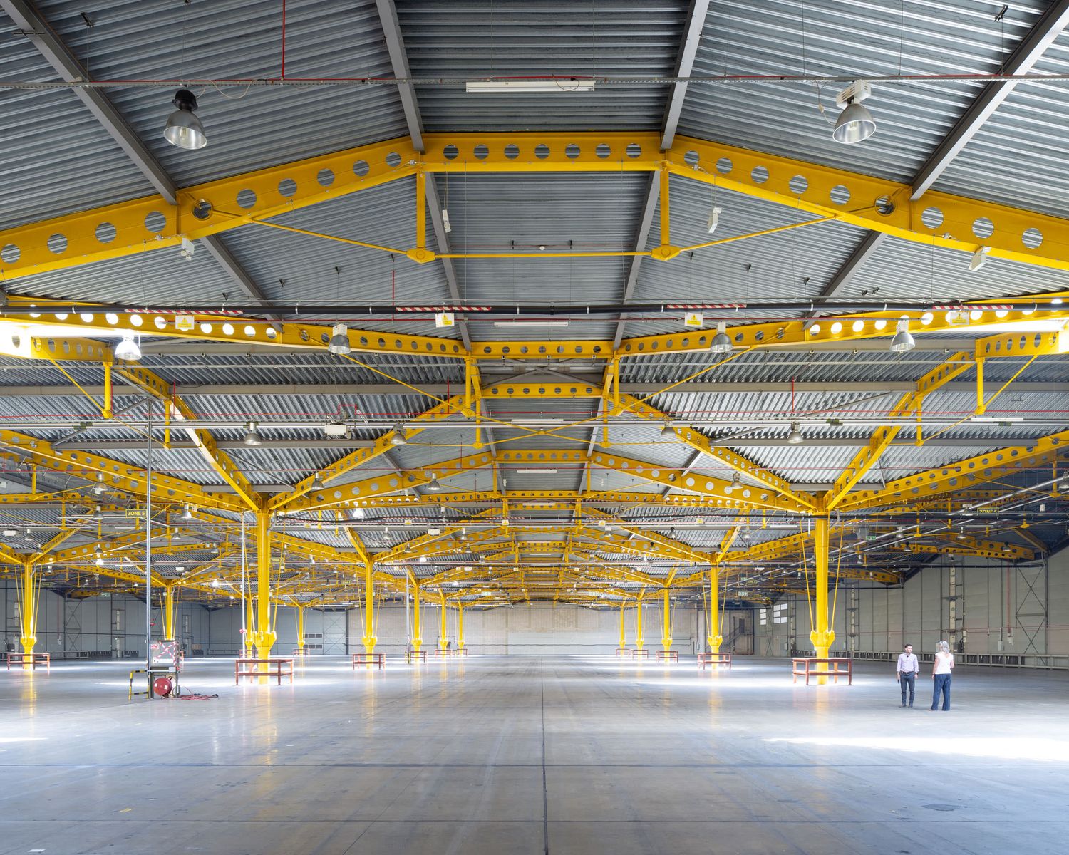 Large empty warehouse interior with yellow steel trusses and beams, polished concrete floor, and two workers in background.