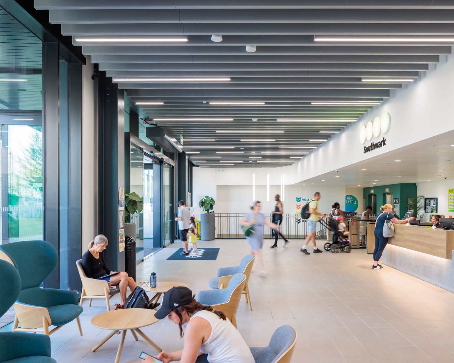 Modern gym lobby with reception desk, seating areas, and people exercising in a bright, open space.