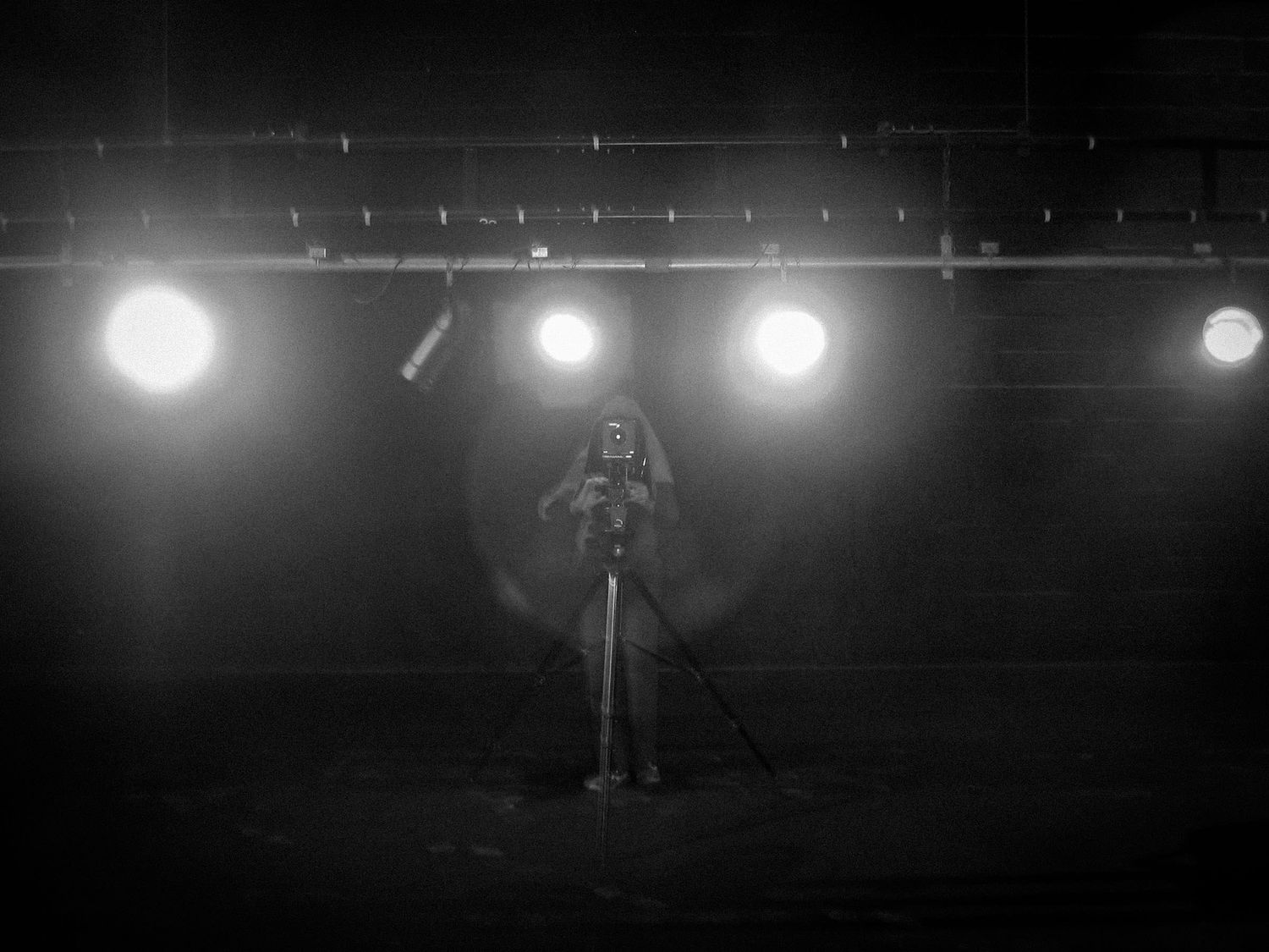 Lone photographer silhouetted against bright stage lights in dark venue.