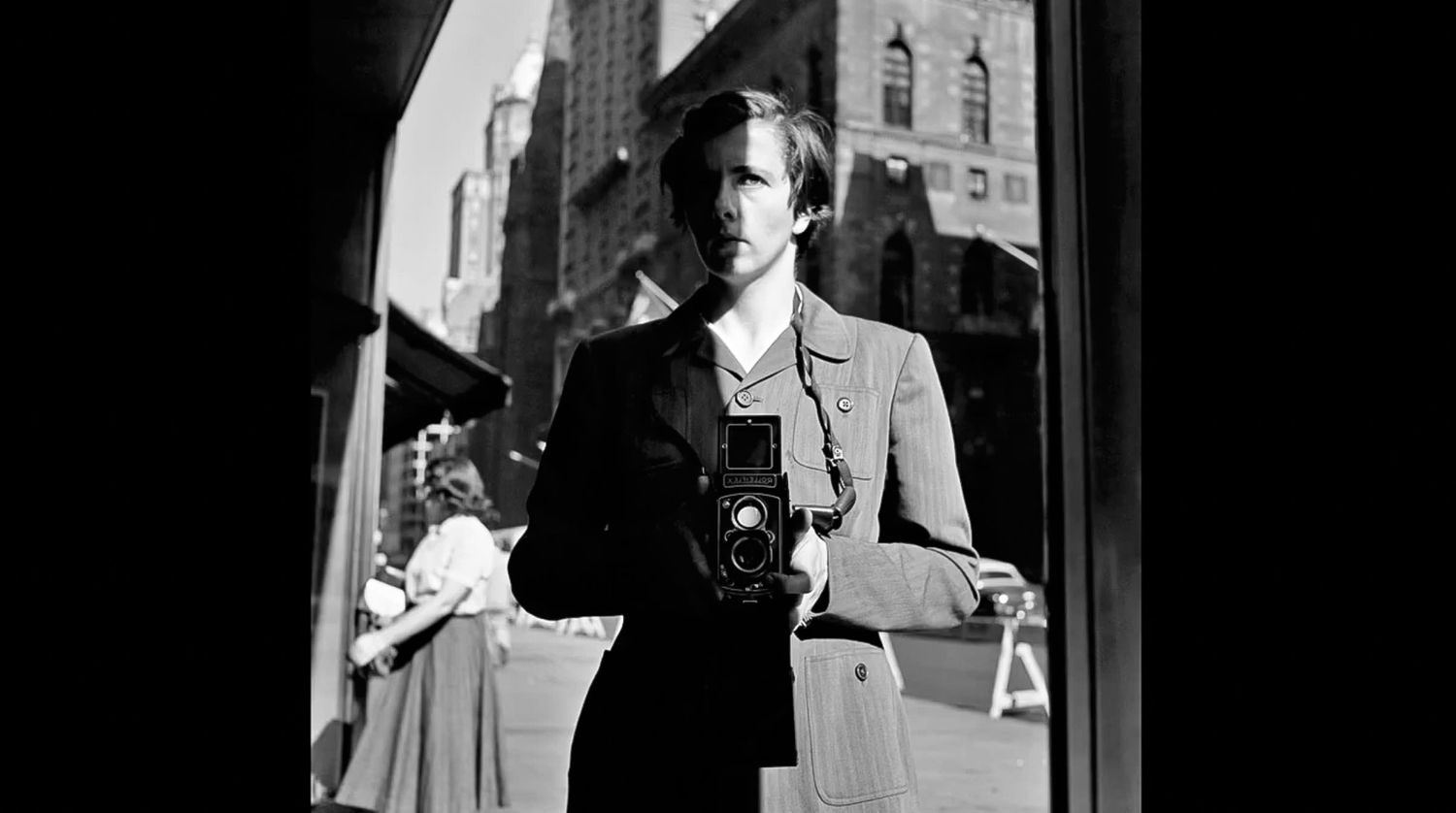 Black and white self-portrait of a person holding a twin-lens reflex camera at chest level, reflected in a window on a city street.