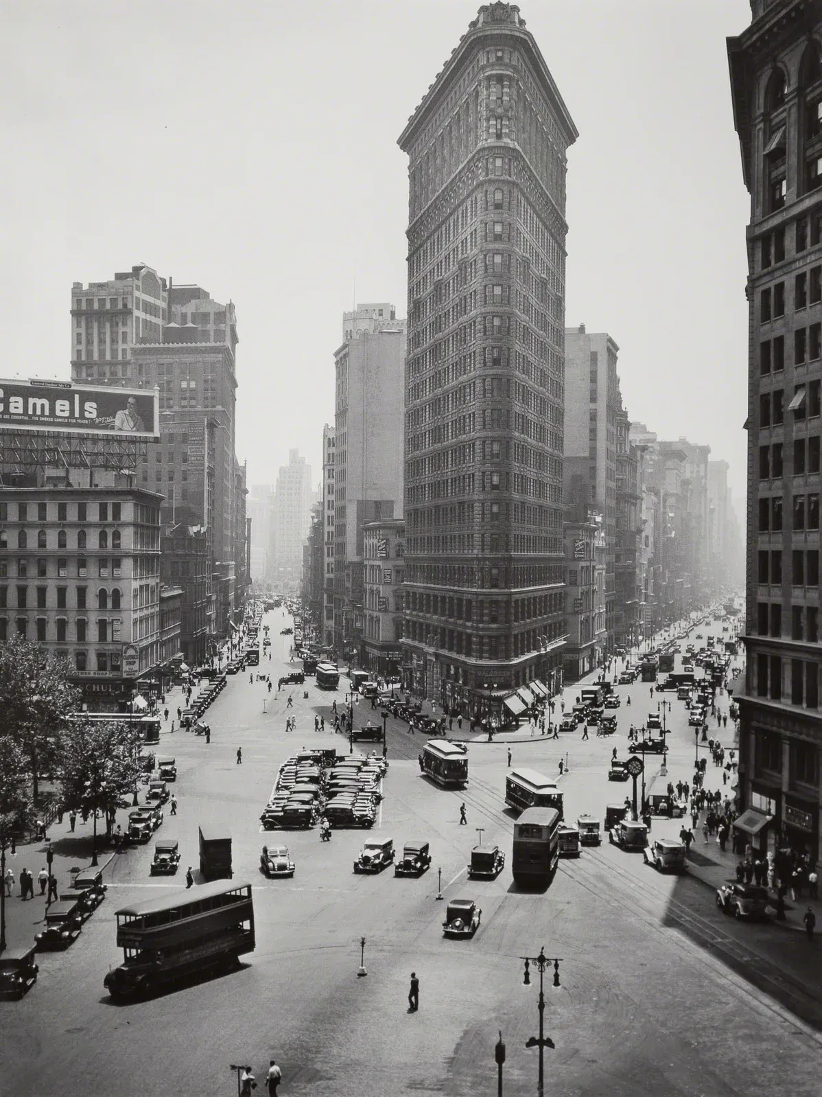 Black and white aerial photo of the Flatiron Building in New York City with vintage cars and busy streets below.