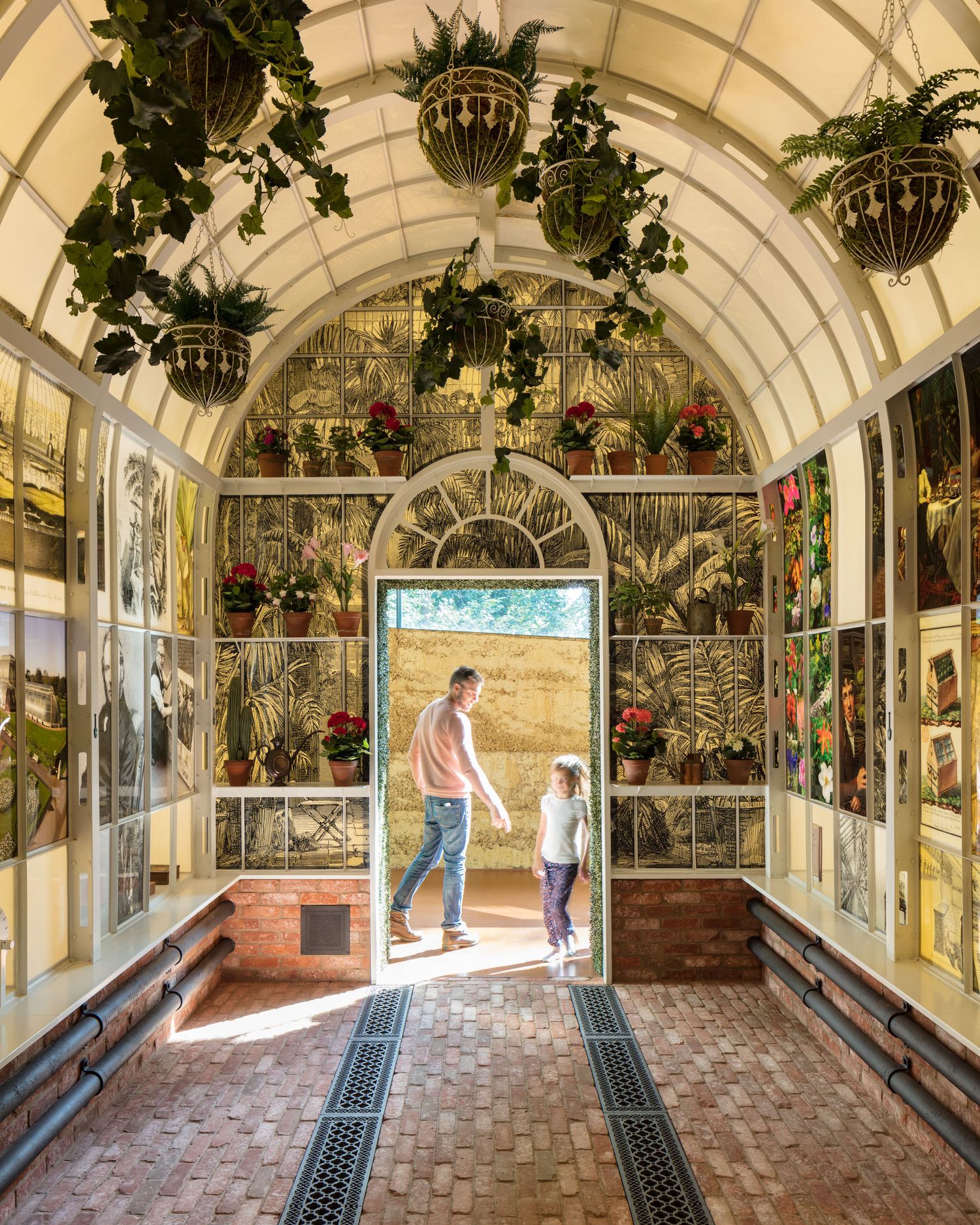 Father and child walk through a Victorian greenhouse corridor with hanging plants and botanical wallpaper.