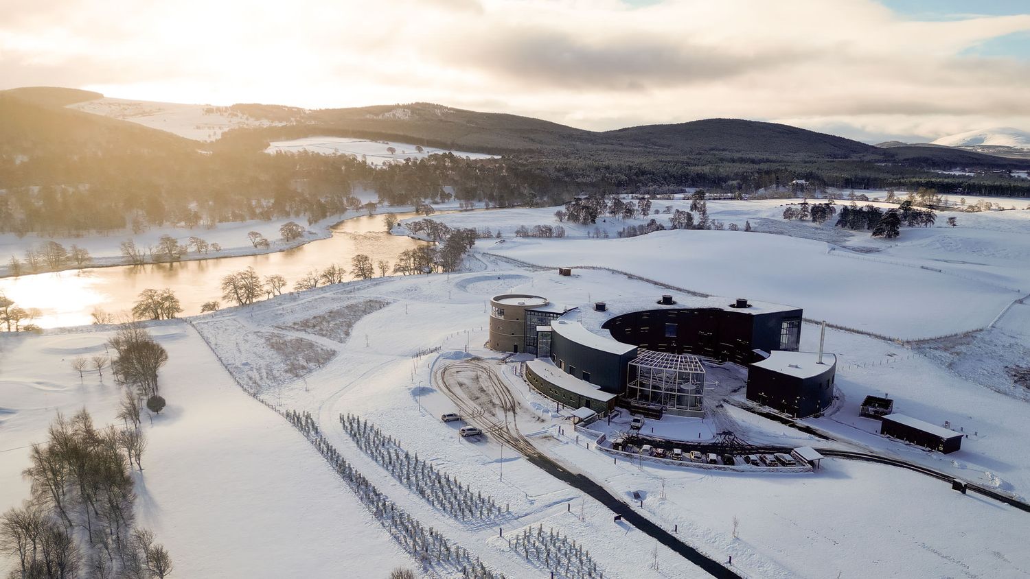 Aerial winter view of a modern circular building surrounded by snow-covered Scottish Highlands landscape at sunset.
