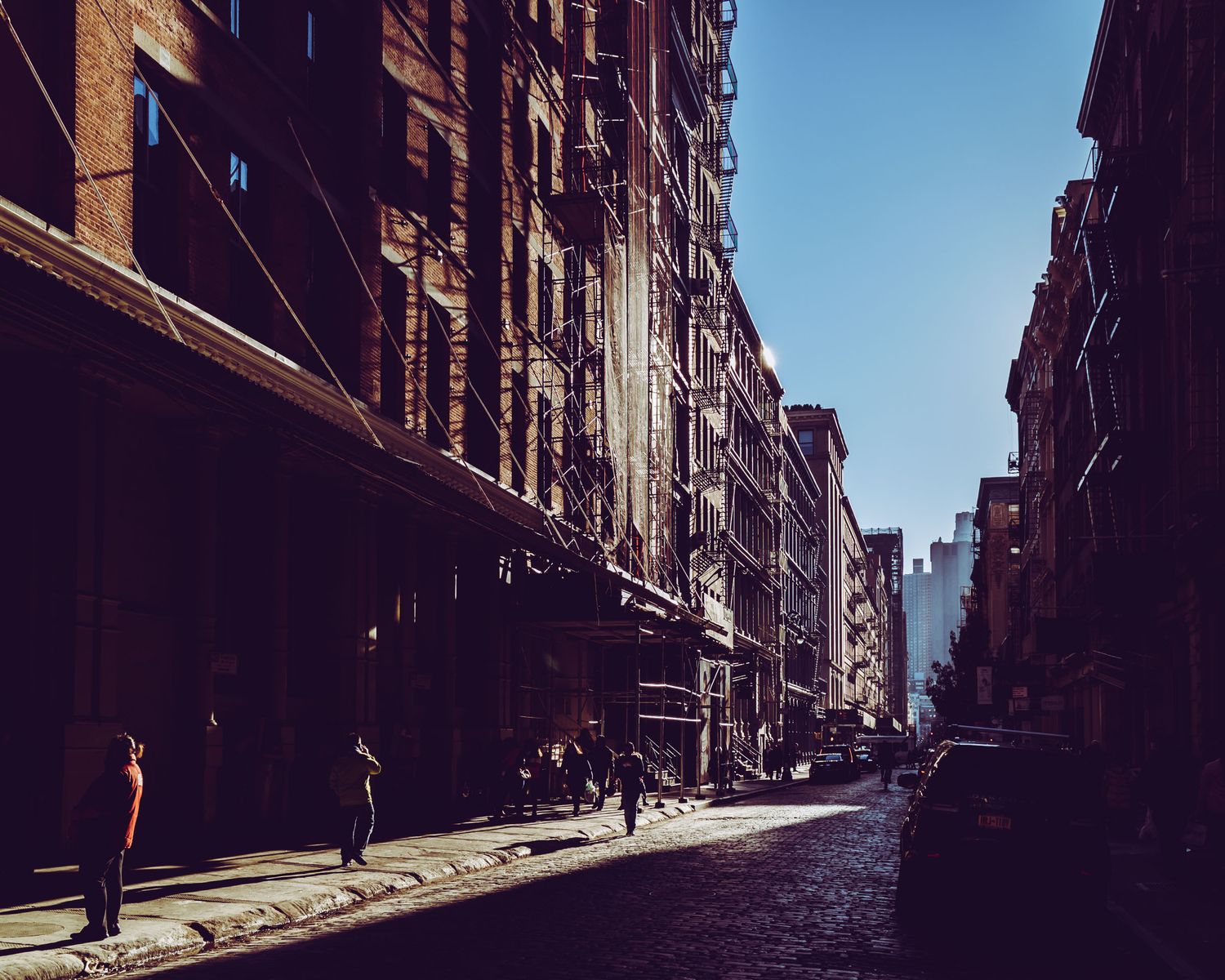 Sunlit urban street lined with historic brick buildings and pedestrians walking on cobblestone sidewalks in a city neighborhood.