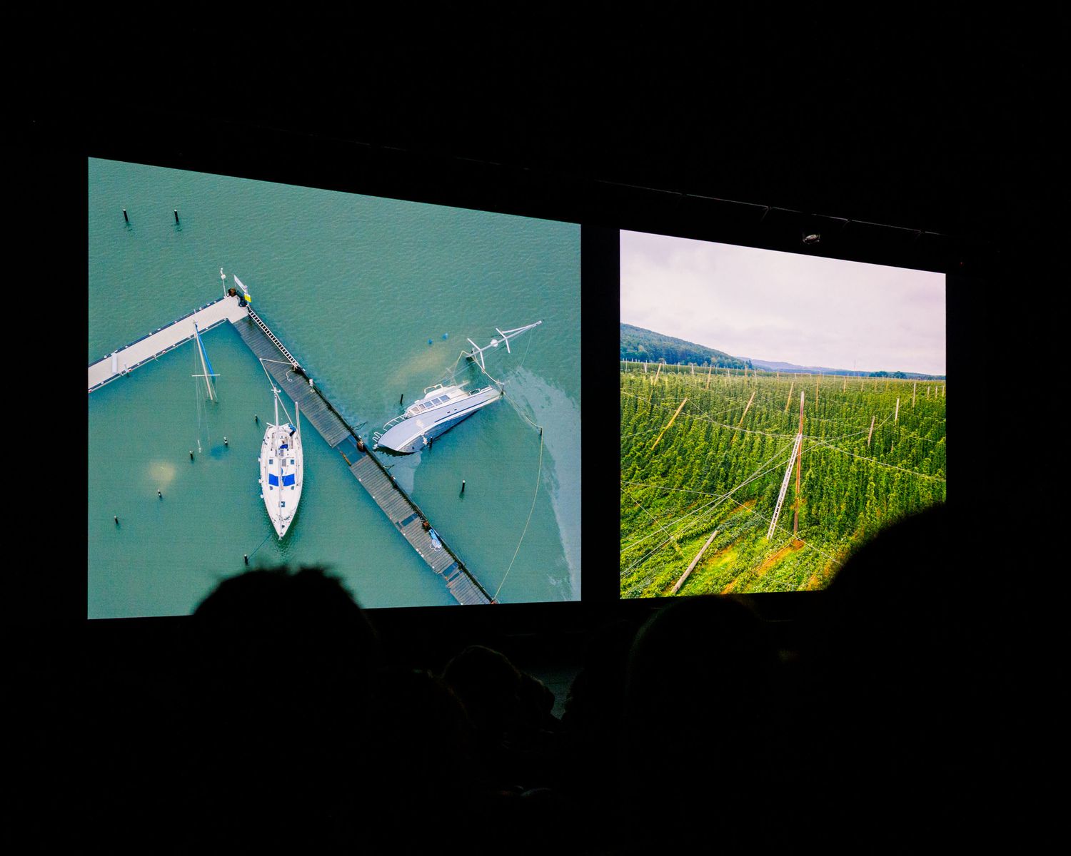 Two projected screens showing aerial sailboat and vineyard landscape images at a dark projection event.