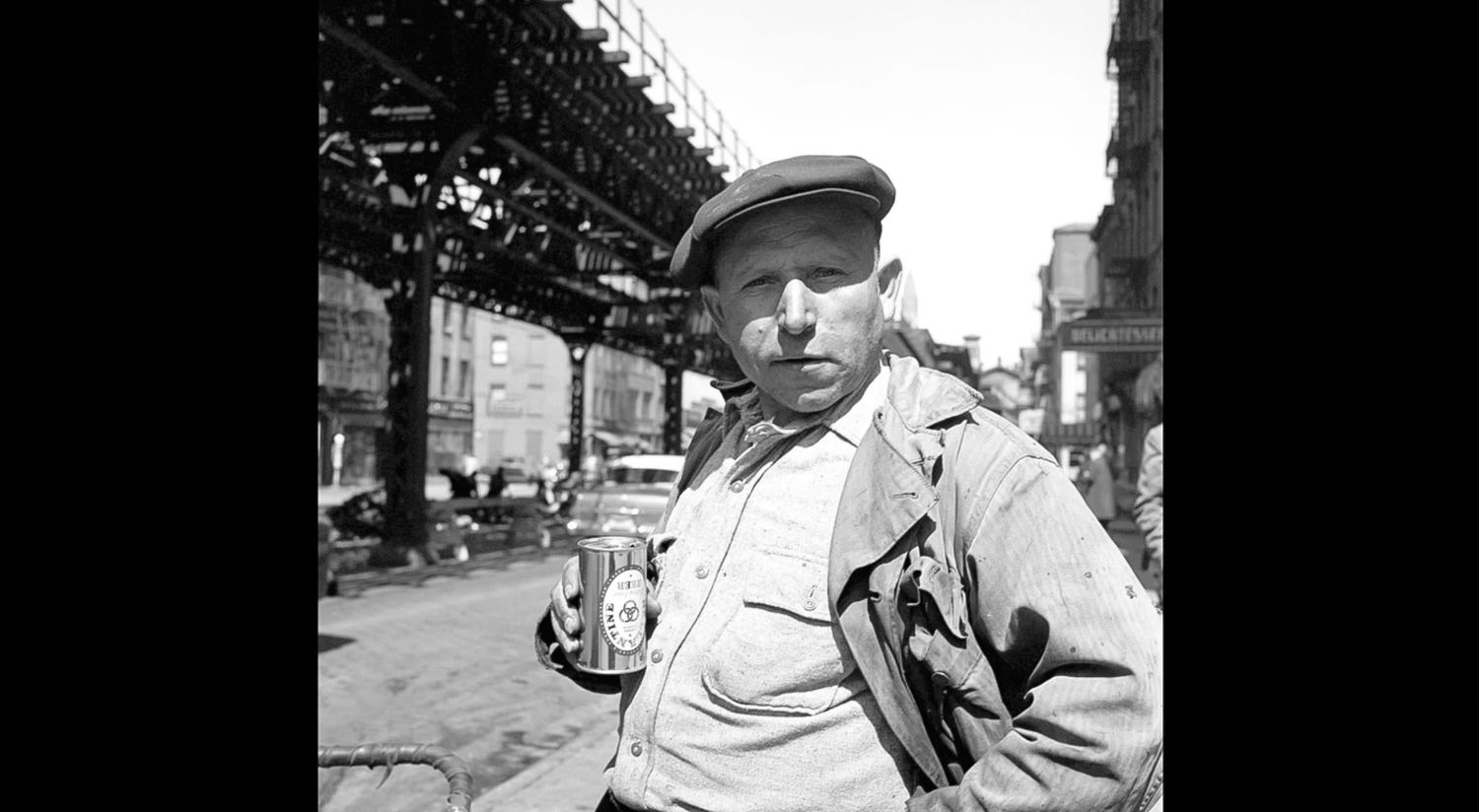 Black and white street photo of a man in a flat cap and jacket holding a can on a city street beneath an elevated train track.