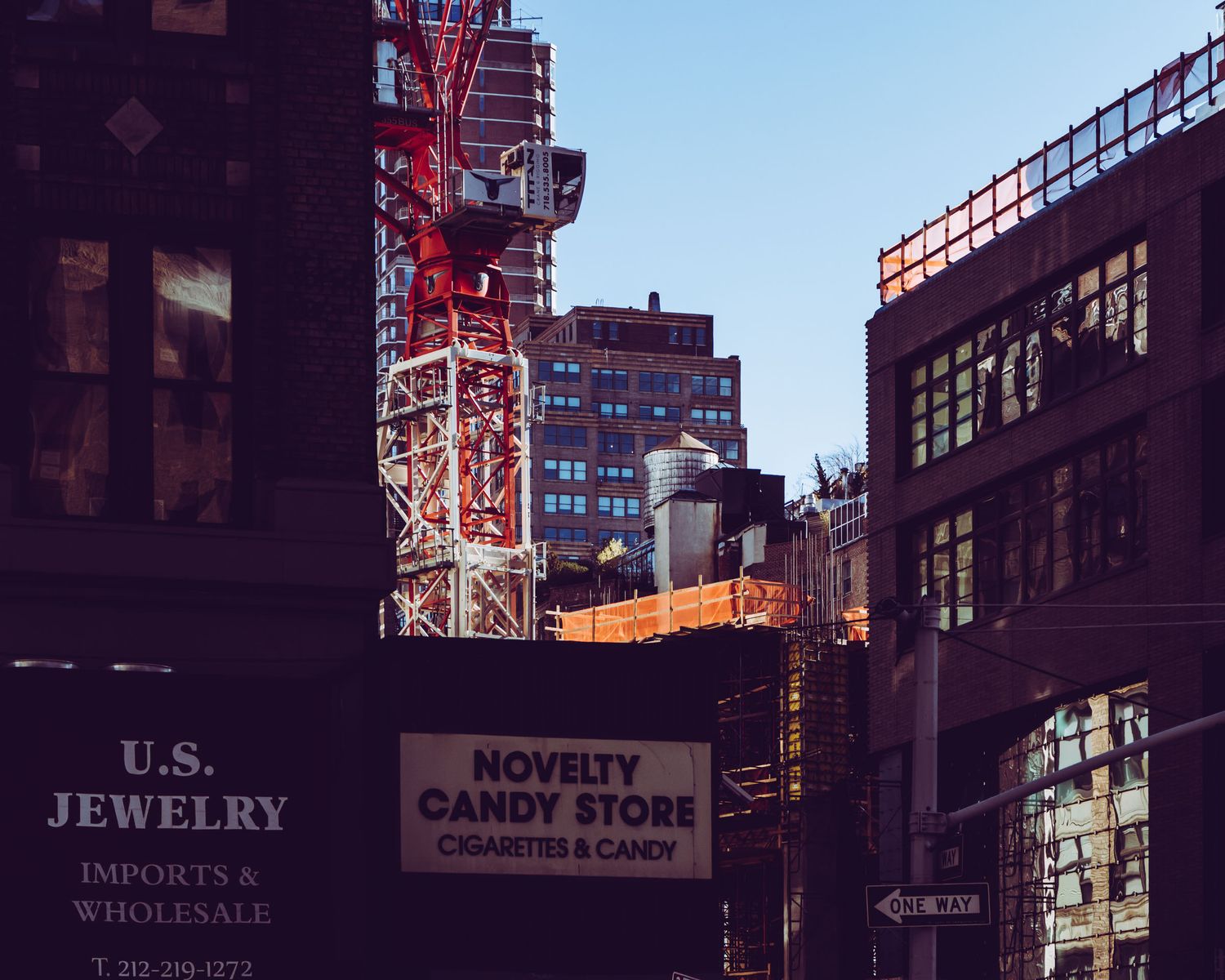 Urban street scene with U.S. Jewelry imports and Novelty Candy Store signs amid NYC buildings and construction crane.
