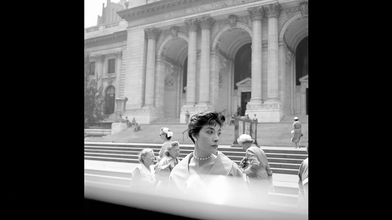 Black and white photo of a short-haired woman standing before a grand columned library building with stone steps and people seated nearby.