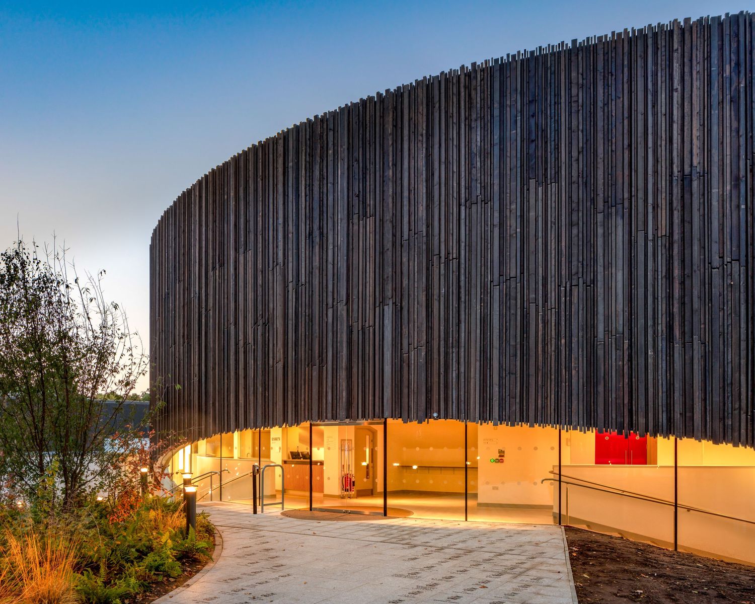 Curved modern building with dark vertical wood cladding facade, illuminated glass entrance at dusk.