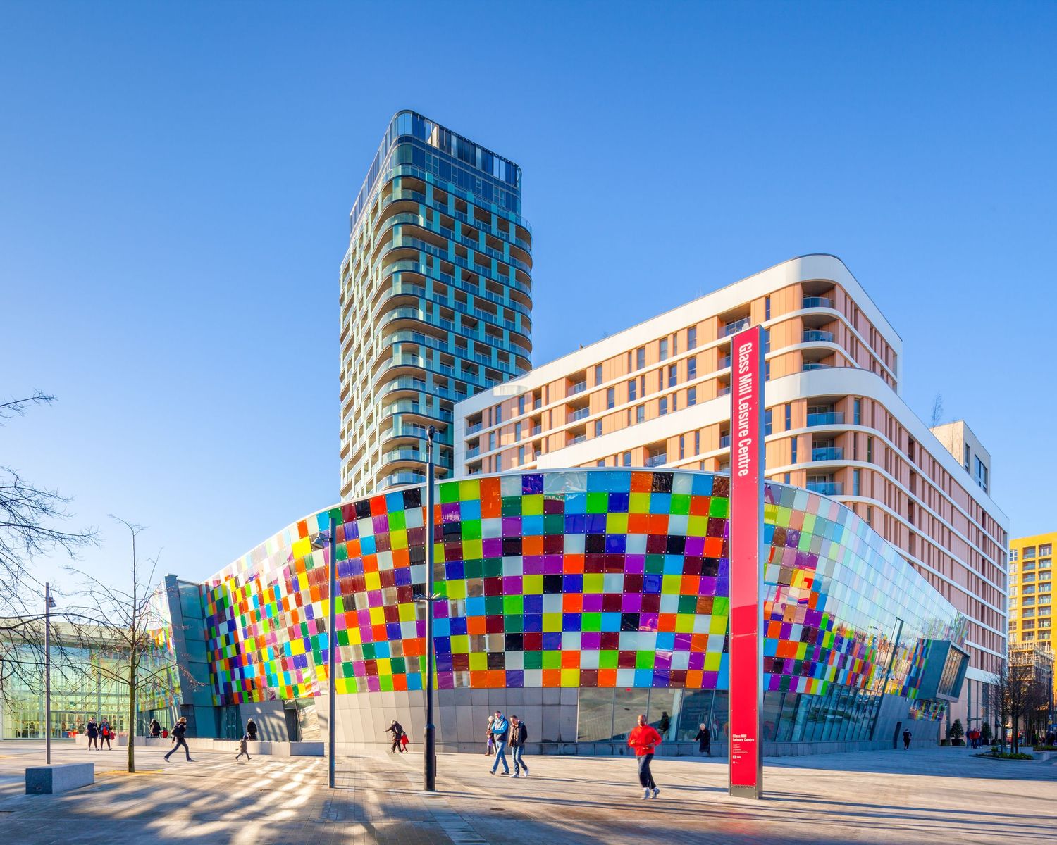 Colorful modern building with vibrant multicolored facade and tall tower under blue sky in urban plaza.