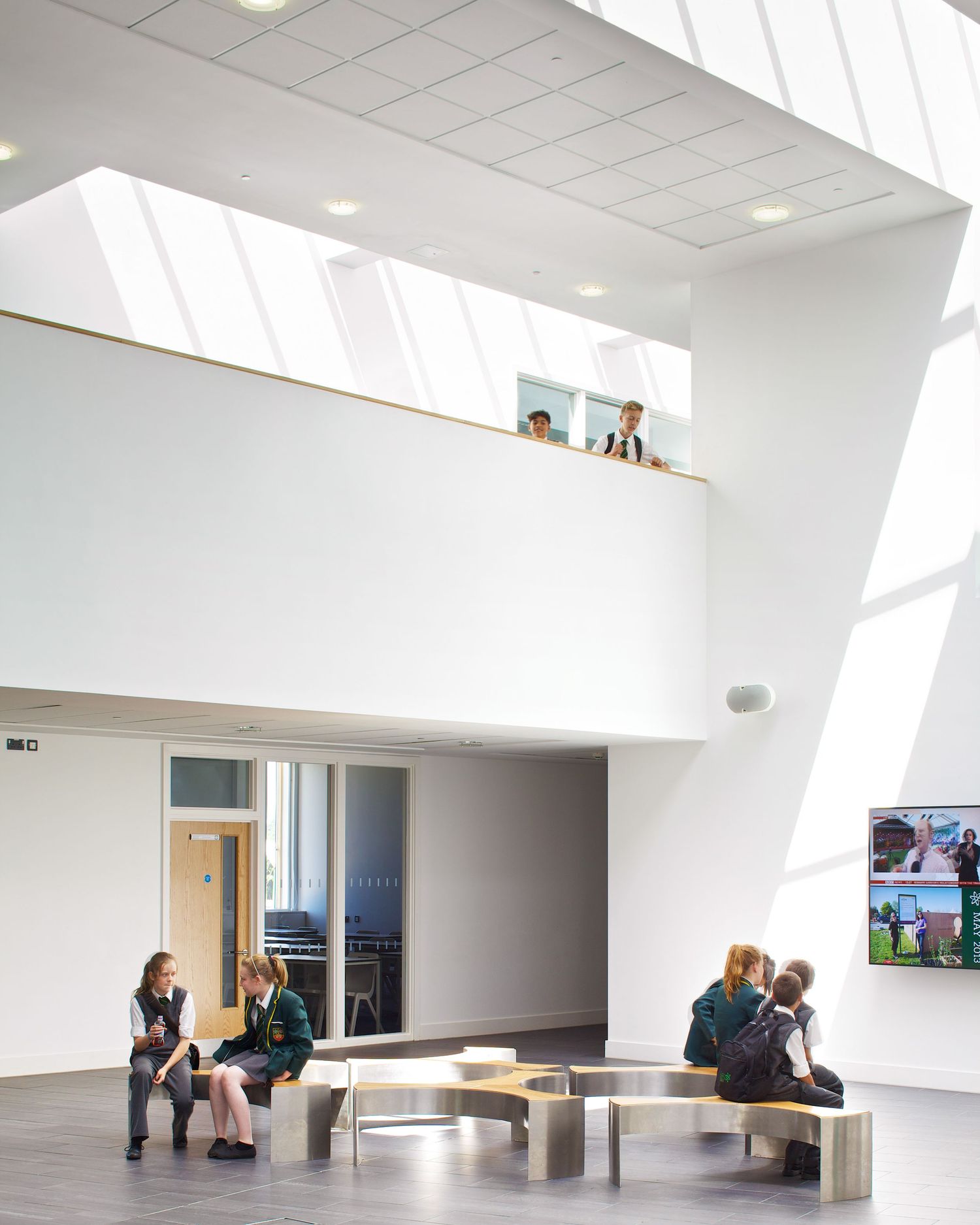 Modern school atrium with students sitting on benches, white walls, skylights, and a mezzanine level above.