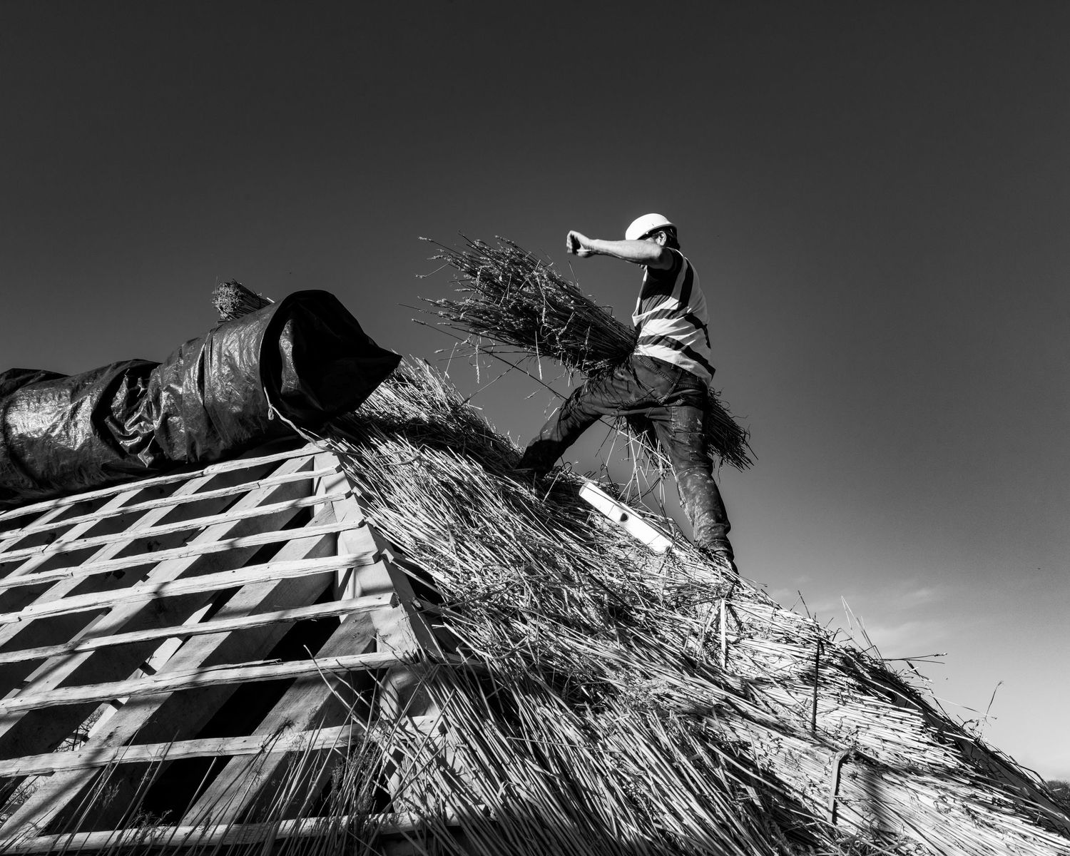 Black and white photo of a worker thatching a roof against a clear sky.