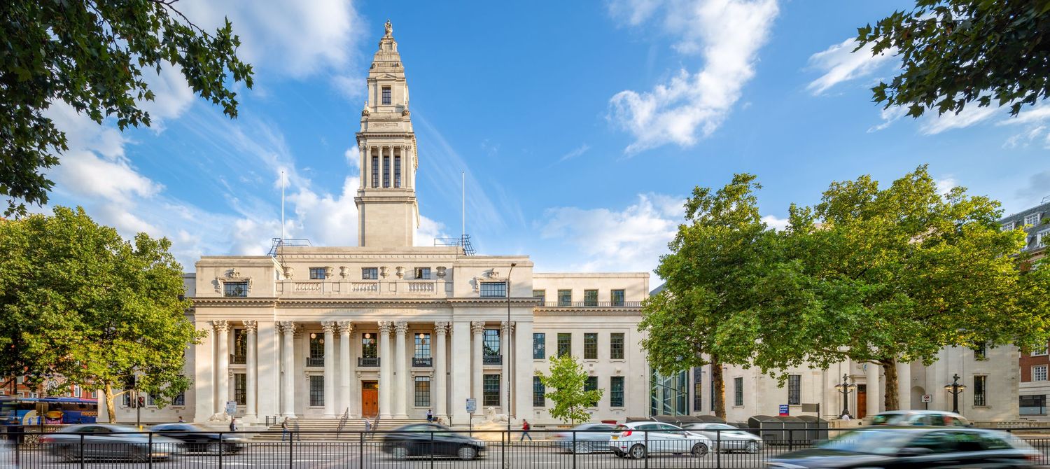 Grand neoclassical government building with clock tower surrounded by lush green trees under a bright blue sky.