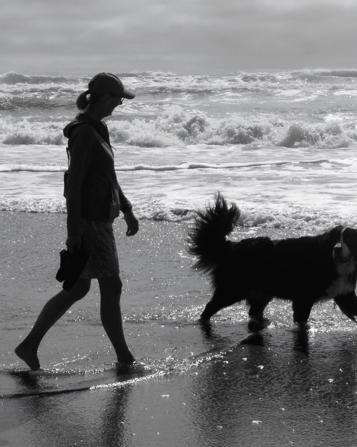 Black and white photo of a woman walking her fluffy dog along a wave-washed beach shoreline.