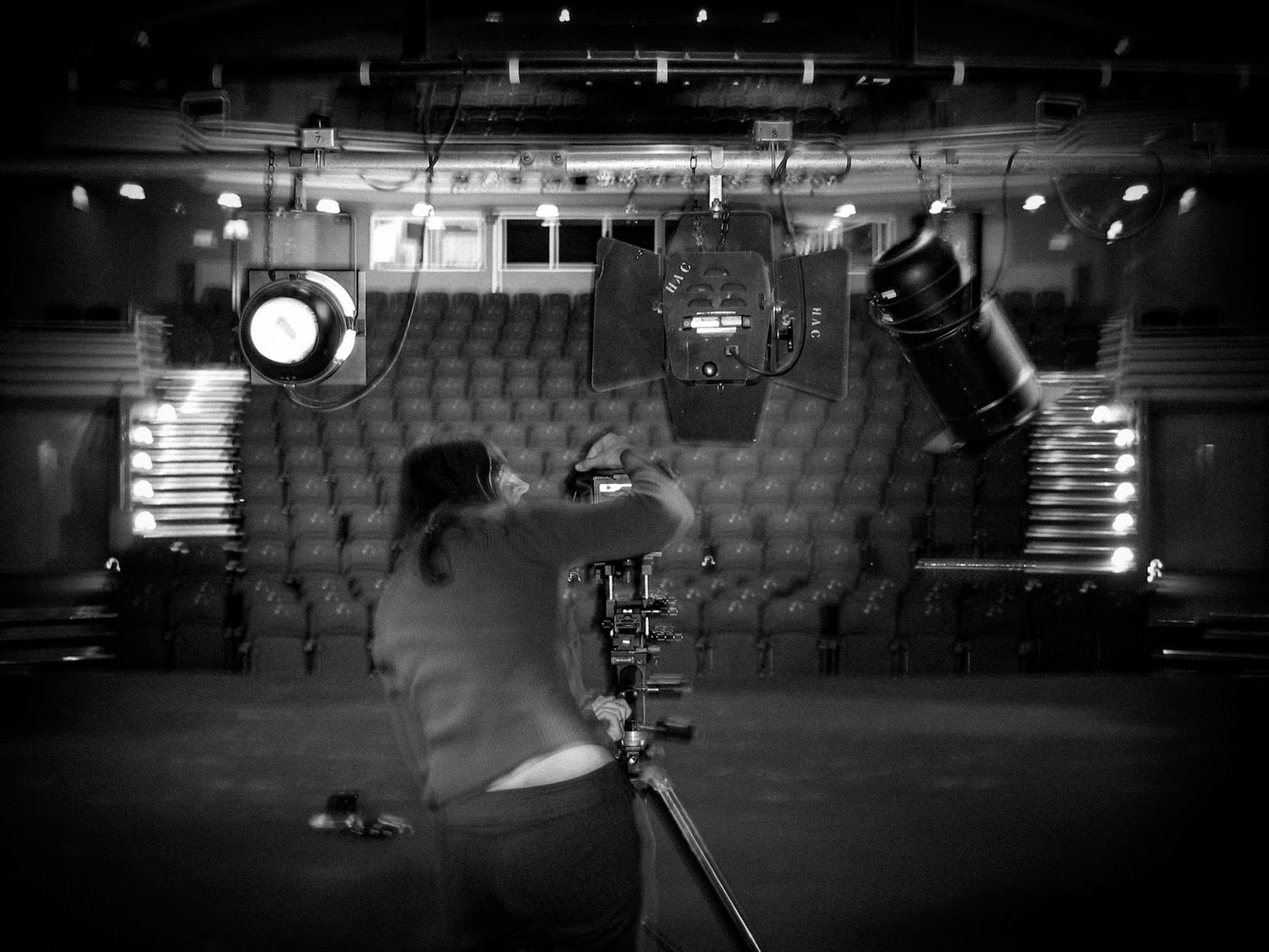 Black and white photo of a cameraman filming an empty theater stage with dramatic stage lighting overhead.