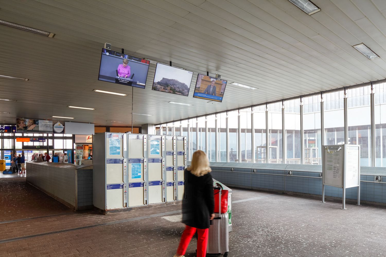 Woman with red luggage standing in a modern airport terminal with information screens and ticket kiosks.