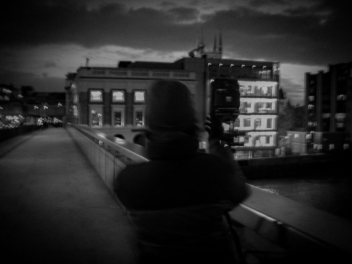 Hooded figure photographing illuminated city buildings from bridge at night.